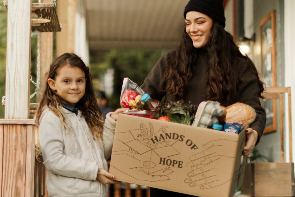 A woman and a girl smile while holding a box labeled "Hands of Hope" filled with groceries, including fruits, vegetables, and packaged food items, on a porch.