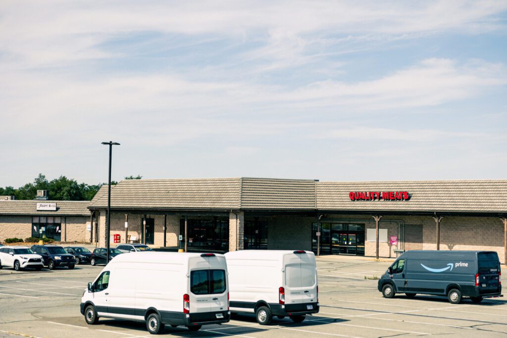 Exterior view of a shopping plaza featuring a "Quality Meats" store, with two white delivery vans parked in the foreground and an Amazon Prime van nearby, under a clear blue sky.