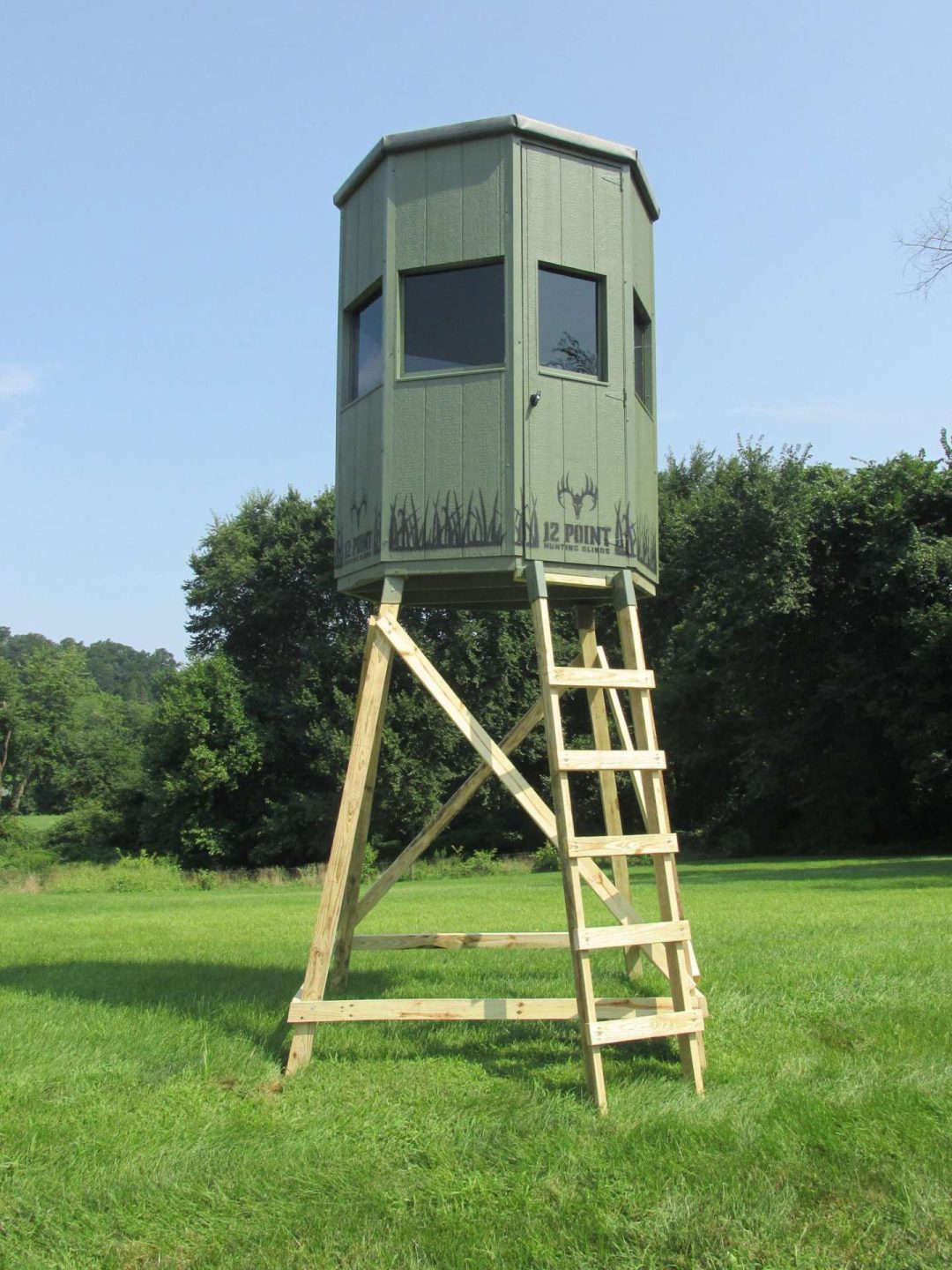 Hunting blind elevated on a wooden platform with a ladder, featuring windows for visibility, set in a green field surrounded by trees. Ideal for deer hunting and wildlife observation.