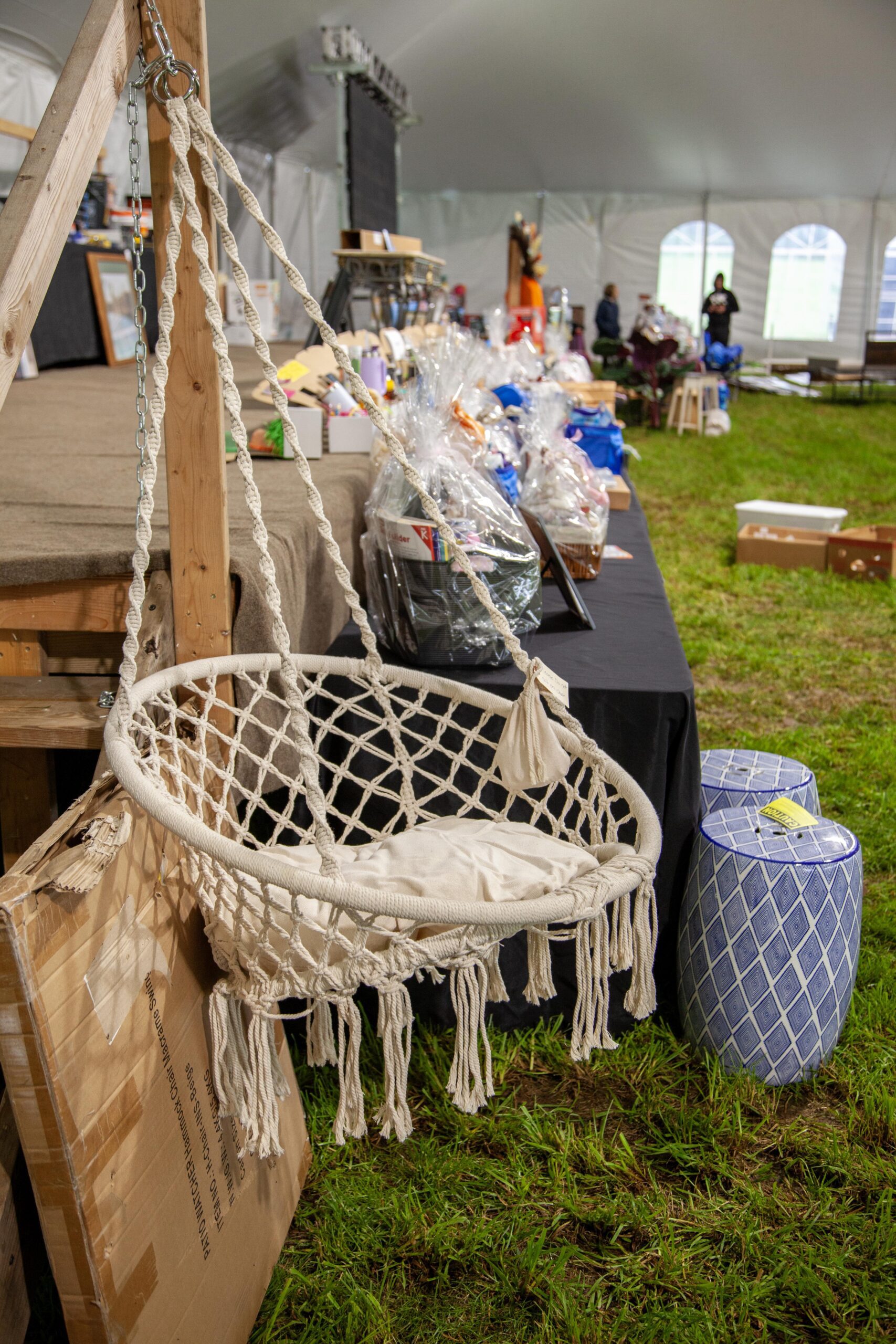 Boho-style hanging chair with cushion displayed at an outdoor event, featuring a backdrop of assorted items for auction on a table, surrounded by grass in a large tent setting.