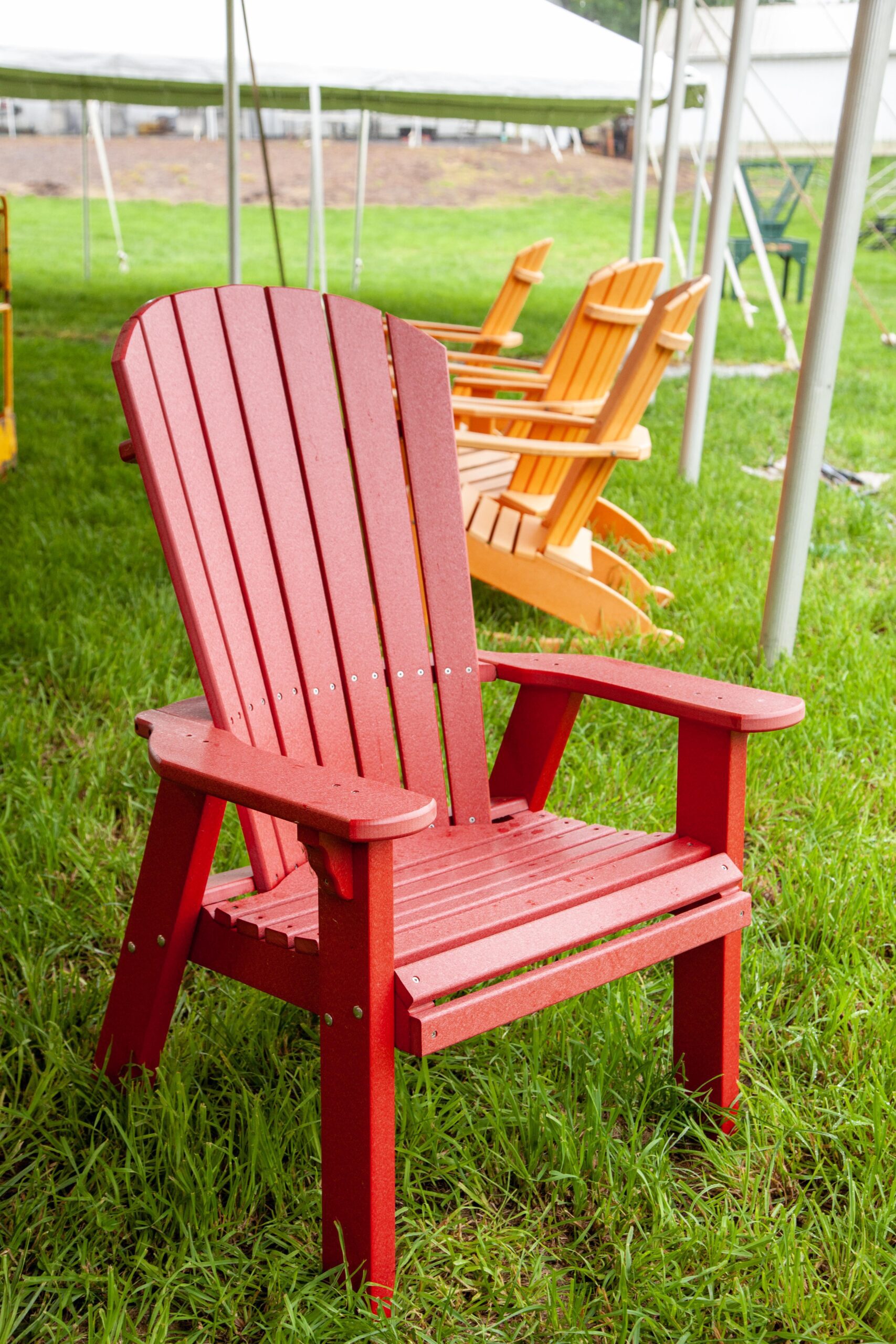 Red Adirondack chair on grass, with additional colorful chairs visible in the background under a tent. Ideal for outdoor relaxation and gatherings.