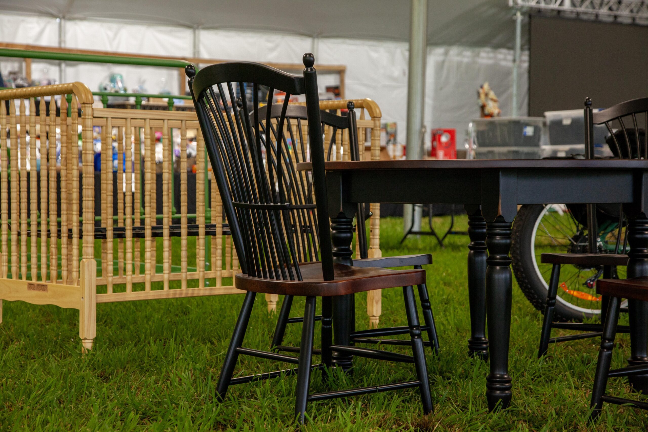 A black wooden chair with a spindle back is positioned next to a dark wood table in a grassy area, with a wooden crib visible in the background under a large tent.