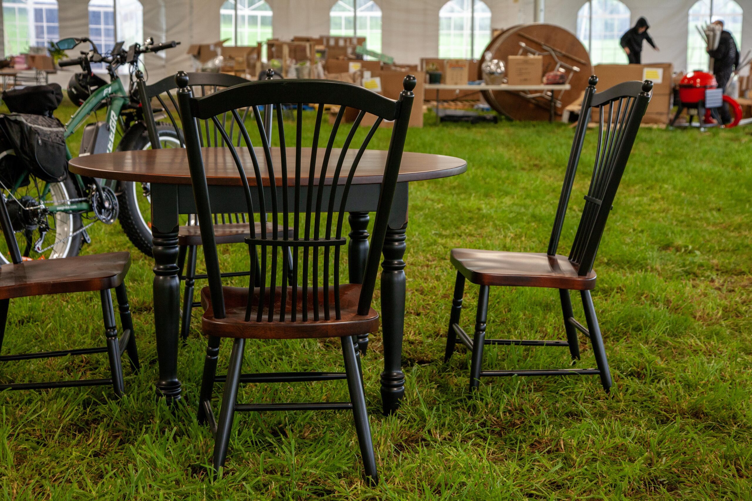 Rustic dining table set with black spindled chairs in a tented outdoor area, surrounded by green grass and cardboard boxes in the background.