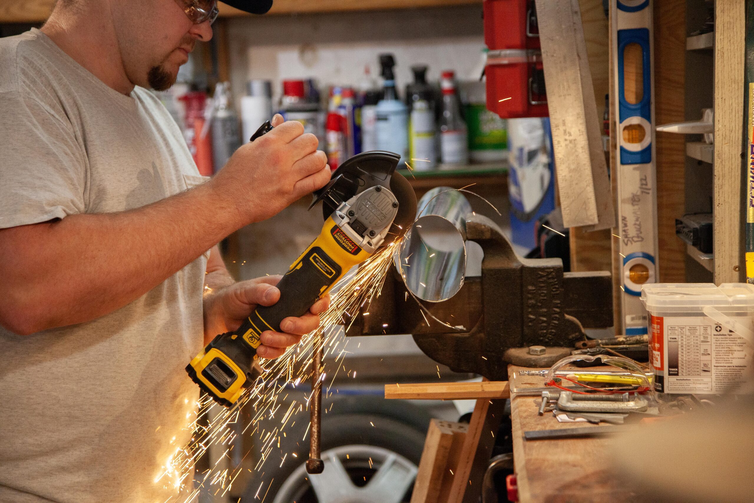 A man wearing safety goggles operates a cordless angle grinder, cutting into a metal pipe. Sparks are flying as he works at a cluttered workbench filled with tools and supplies, showcasing a hands-on metalworking project in a garage workshop.