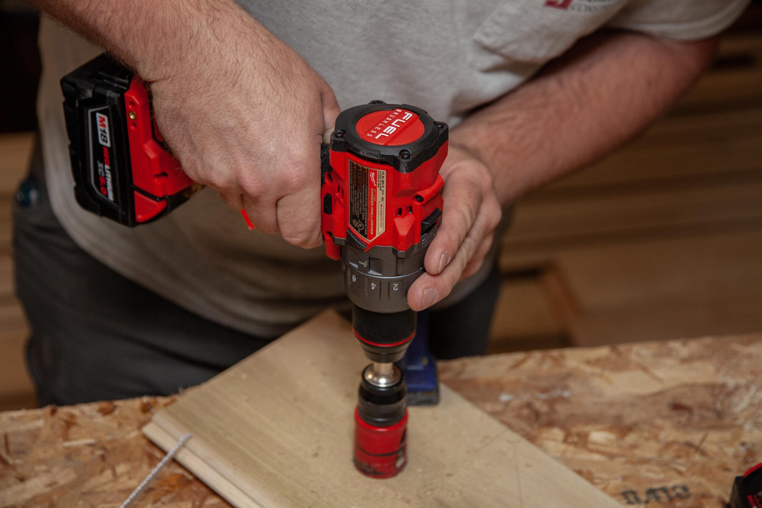 Close-up of a person using a Milwaukee cordless drill with a battery pack to fasten screws into a wooden surface, showcasing a hands-on construction or DIY project in a workshop setting.
