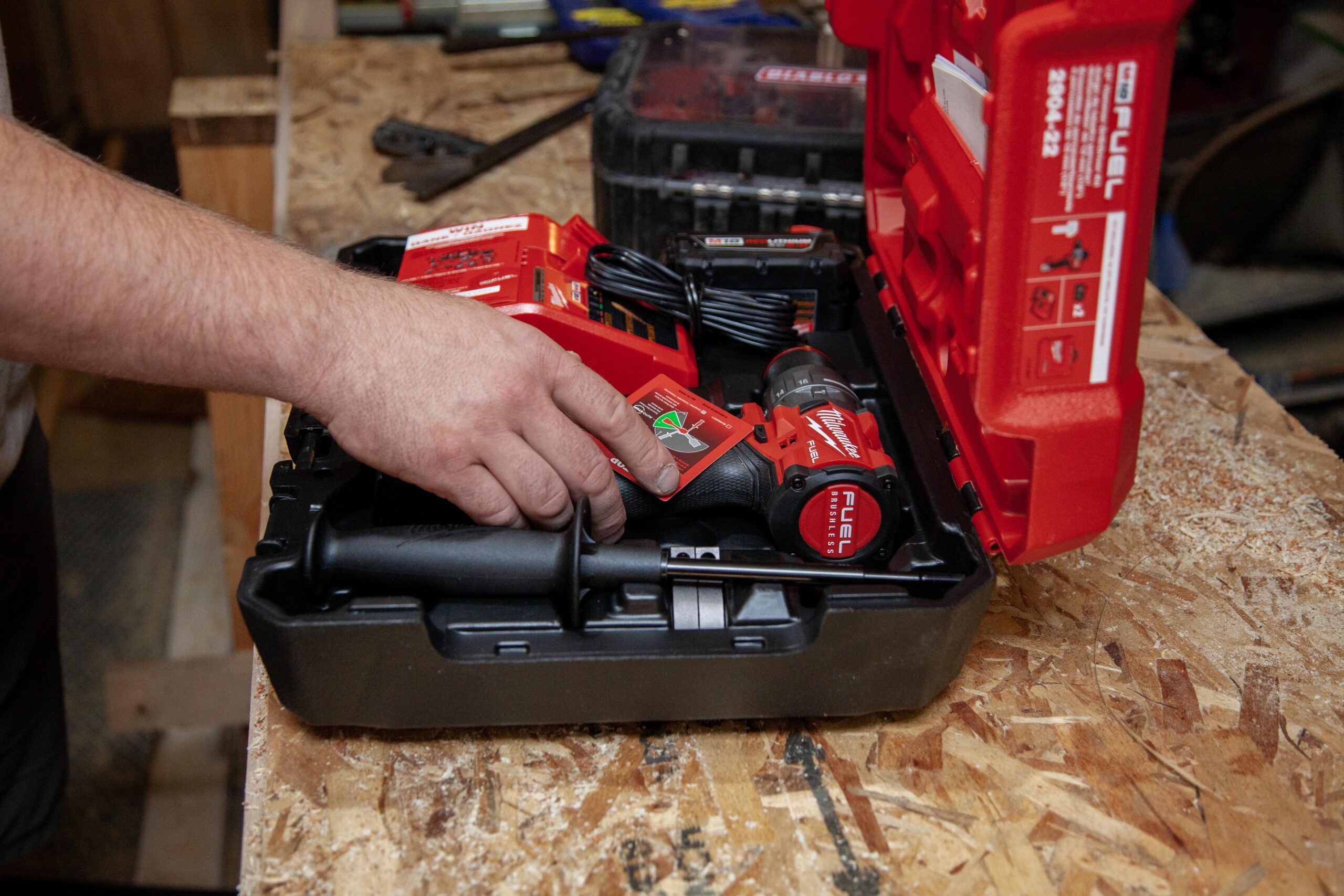 Hand reaching for a Milwaukee Fuel power tool inside a red carrying case, showcasing batteries and chargers on a wooden workbench.