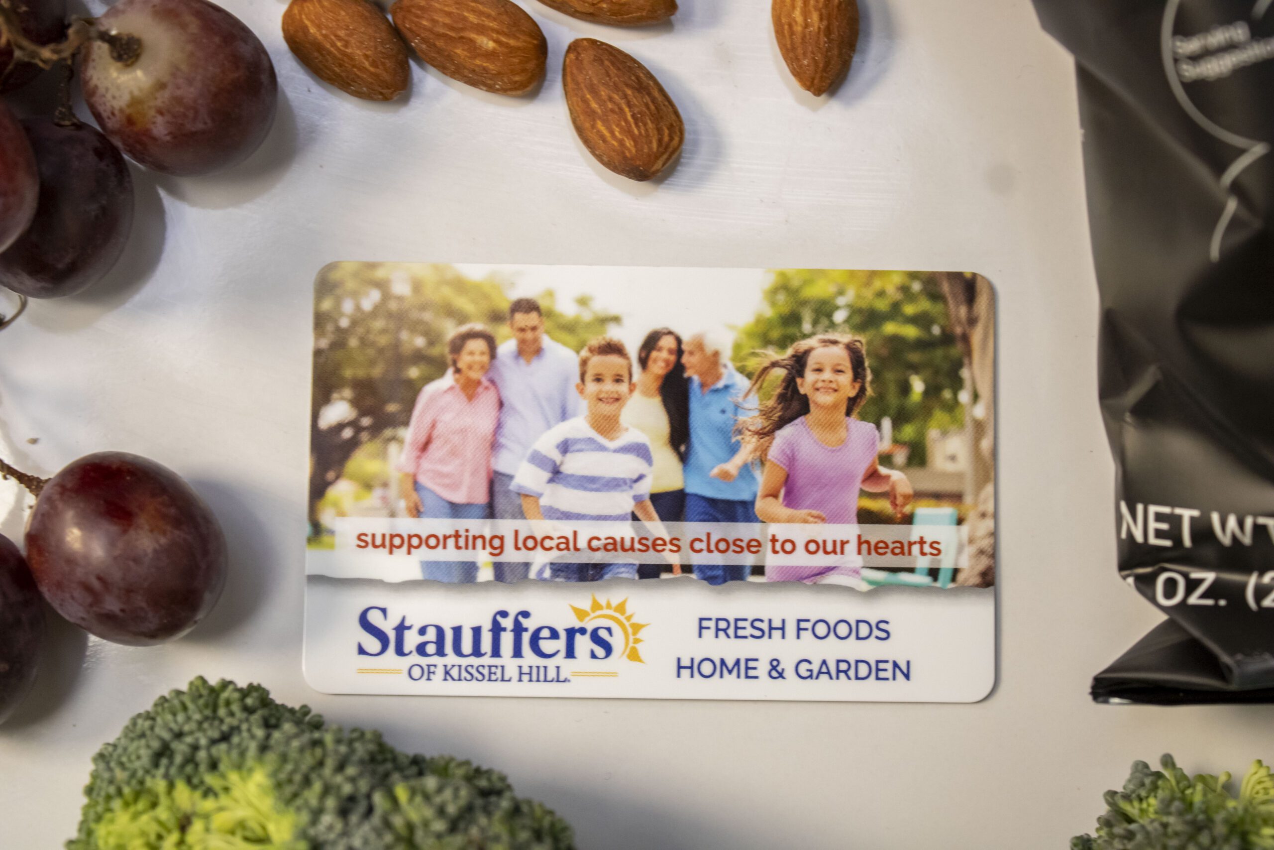 Stauffers card with a family, grapes, almonds, and broccoli, promoting local causes and fresh foods.