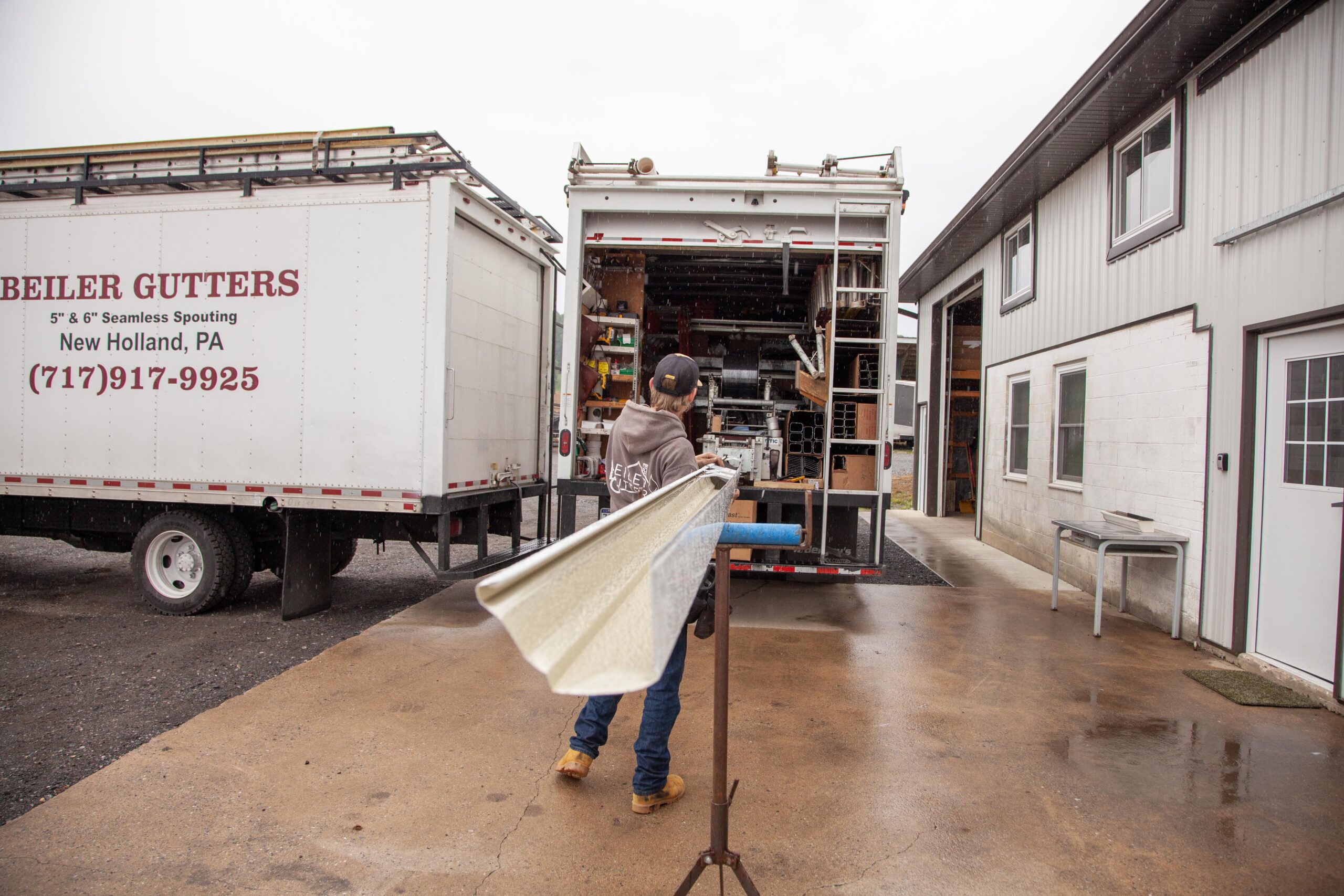 Employee handling gutter materials outside a workshop, with Beiler Gutters trucks and equipment visible in the background, emphasizing seamless spouting services in New Holland, PA.