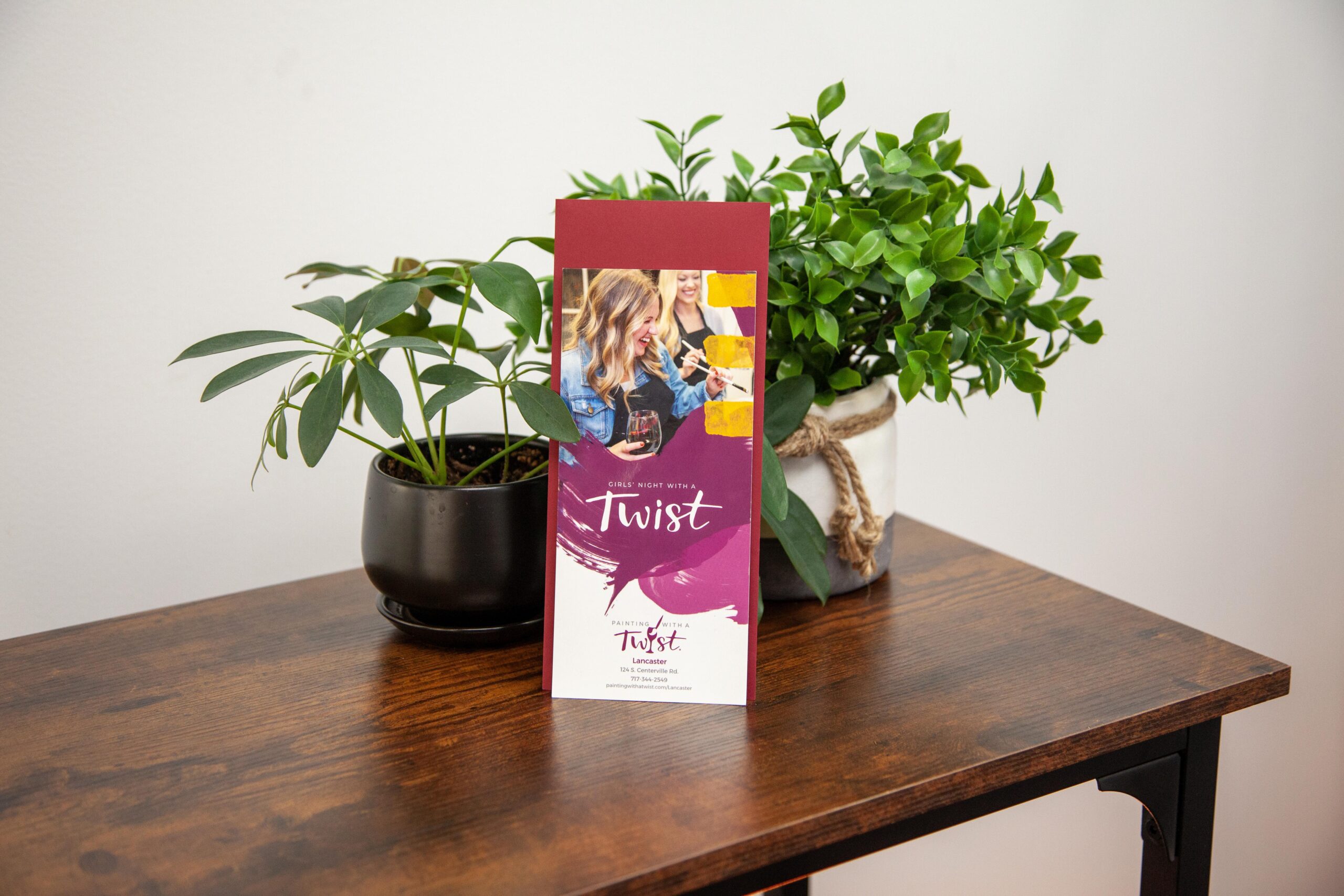 Promotional brochure for "Girls Night with a Twist" event, featuring vibrant colors and images, placed on a wooden table next to a decorative plant.