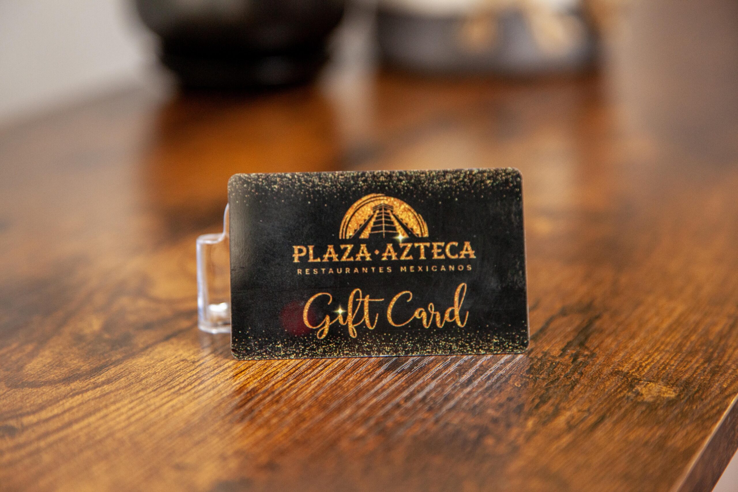 Plaza Azteca Mexican Restaurant gift card displayed on a wooden table, featuring a sleek black design with gold accents and text indicating it's a gift card.