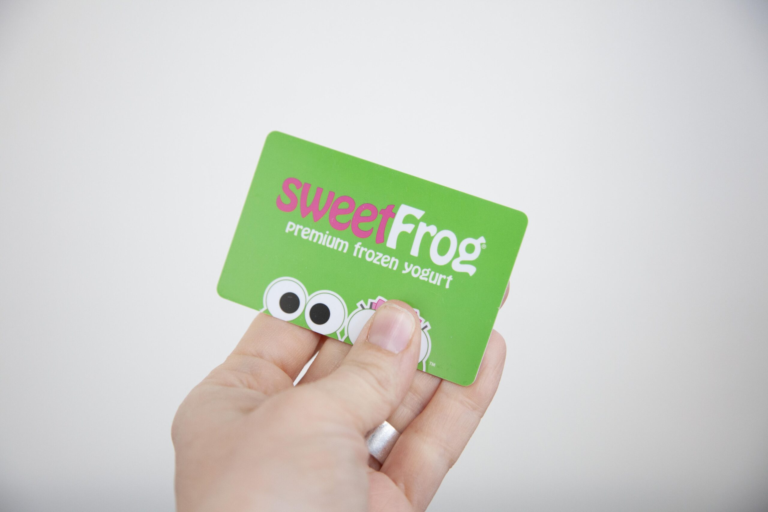 Hand holding a SweetFrog premium frozen yogurt loyalty card, featuring a bright green background with playful eyes and logo, highlighting the brand's focus on delicious frozen yogurt treats.