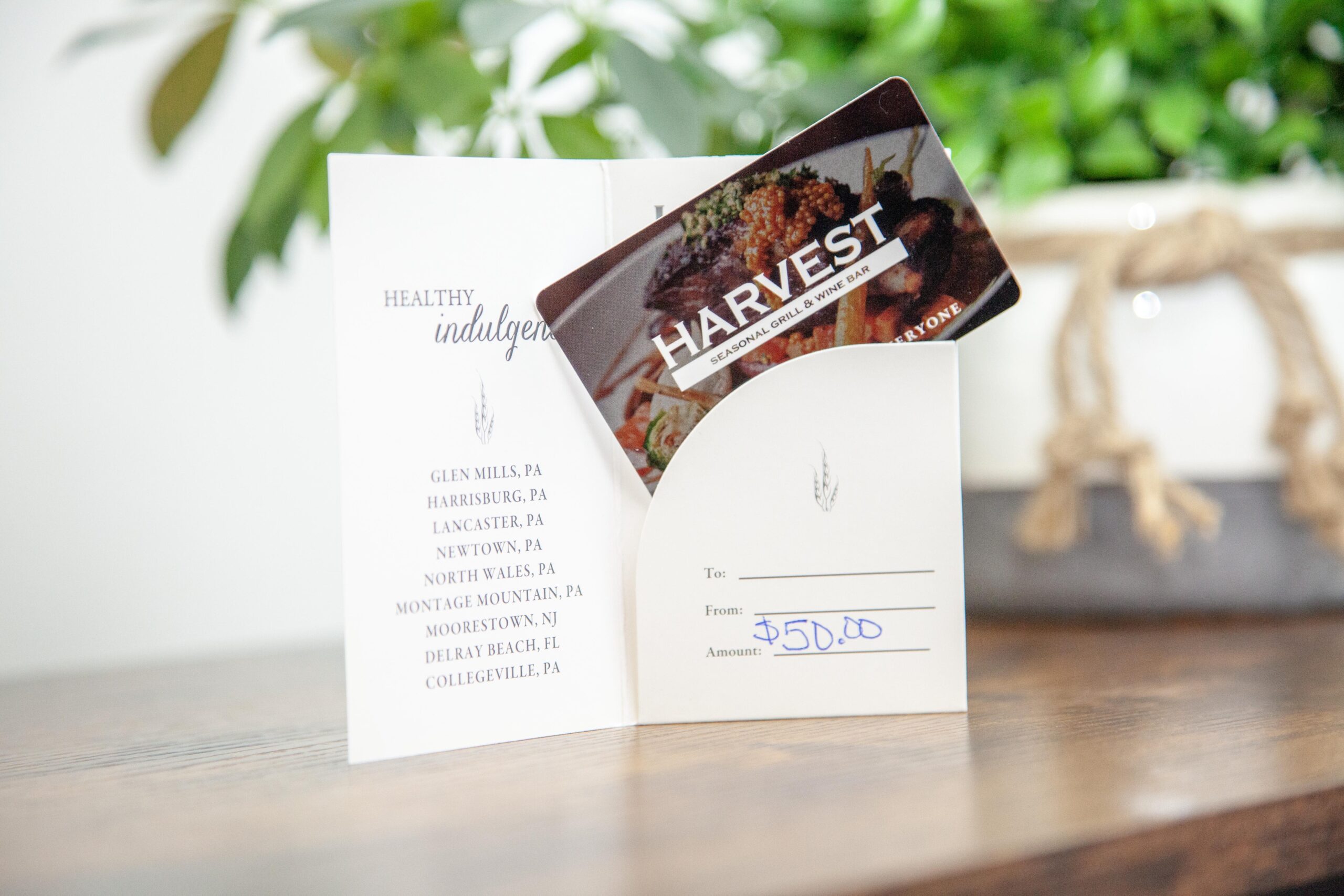 Gift card for Harvest Seasonal Grill & Wine Bar displayed in a decorative envelope. The card is valued at $50 and includes a list of locations such as Glen Mills, Harrisburg, and Delray Beach. Ideal for dining or gifting experiences at a farm-to-table restaurant.