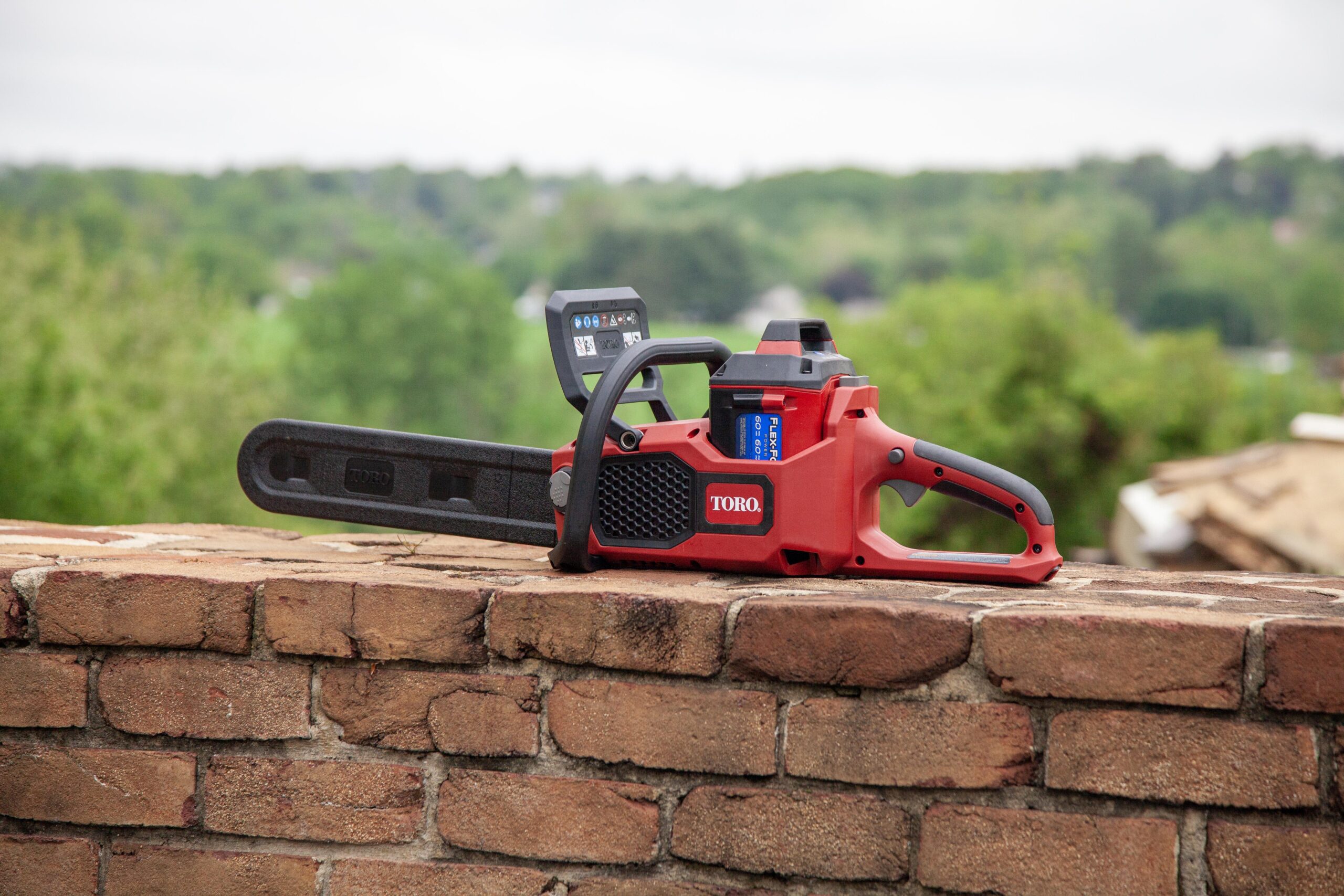 Toro cordless chainsaw resting on a brick wall, showcasing its sleek design and a digital display. The background features a lush green landscape, indicating outdoor usage. Ideal for homeowners and landscaping tasks.