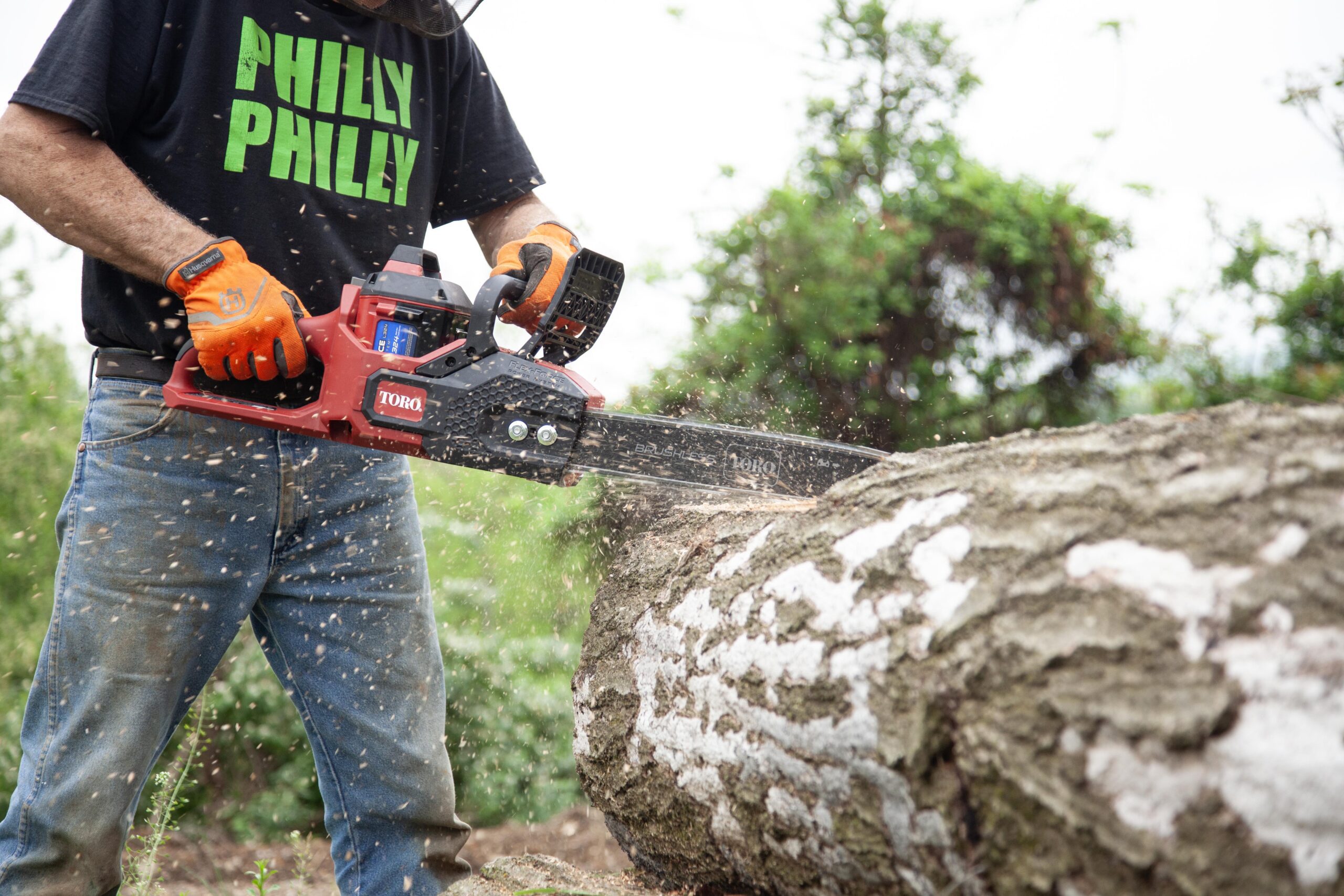 A person using a Toro chainsaw to cut a log, with wood chips flying in the air, wearing gloves and a black shirt.