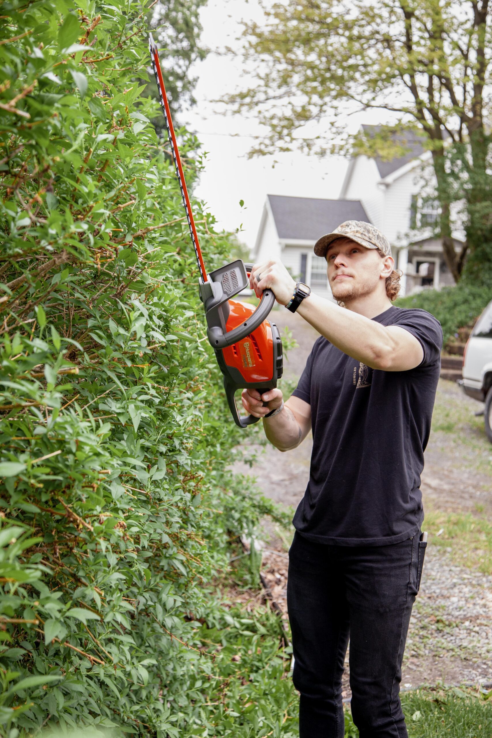 Man using an electric hedge trimmer to shape bushes in a residential yard, wearing a black t-shirt and a camouflage cap.