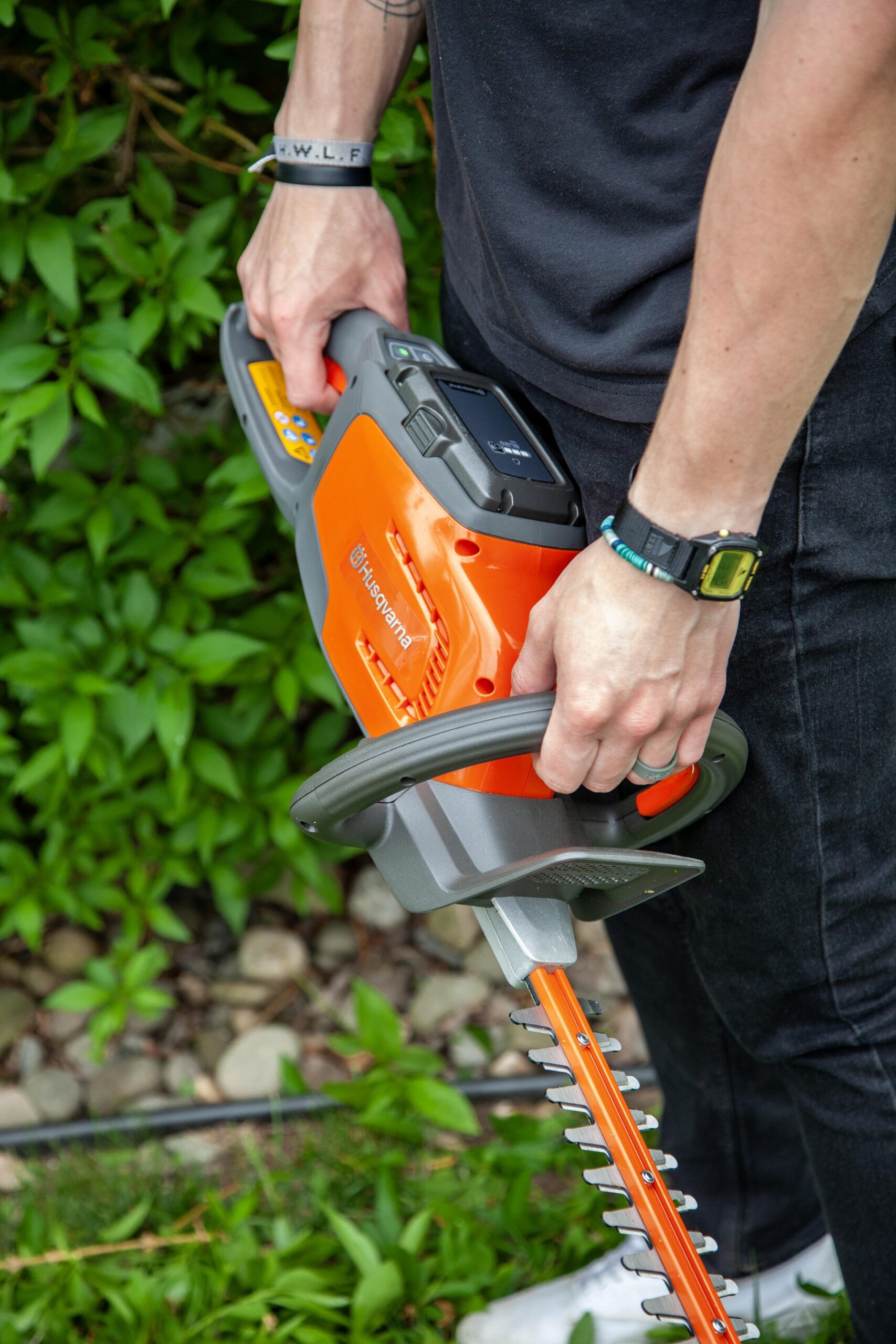 A person holds a Husqvarna battery-operated hedge trimmer, demonstrating its ergonomic design and user-friendly controls, surrounded by green foliage and garden stones.
