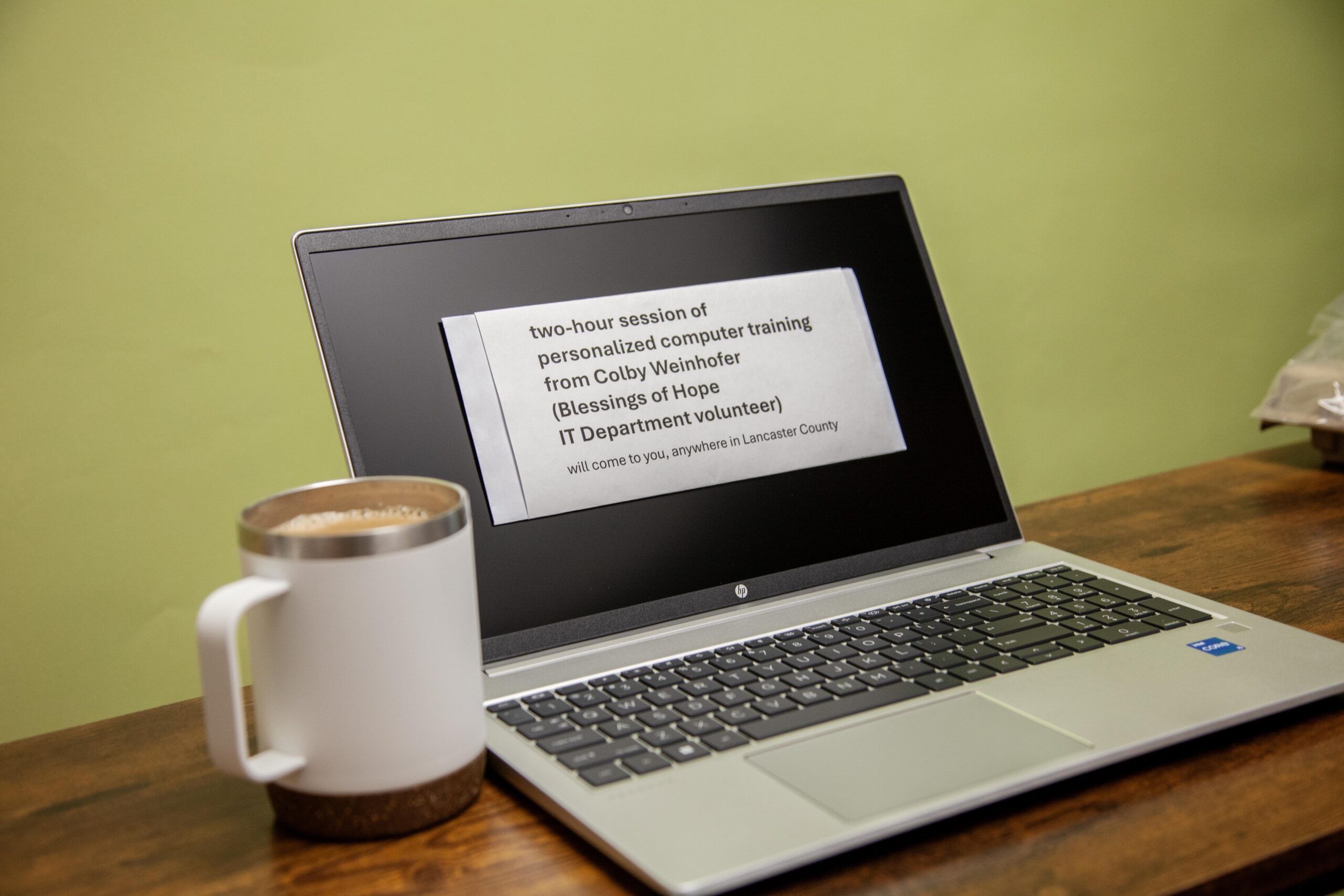 Laptop displaying a flyer for a two-hour personalized computer training session by Colby Weinhofer, a volunteer from Blessings of Hope, offered in Lancaster County. A coffee mug is placed next to the laptop on a wooden table.