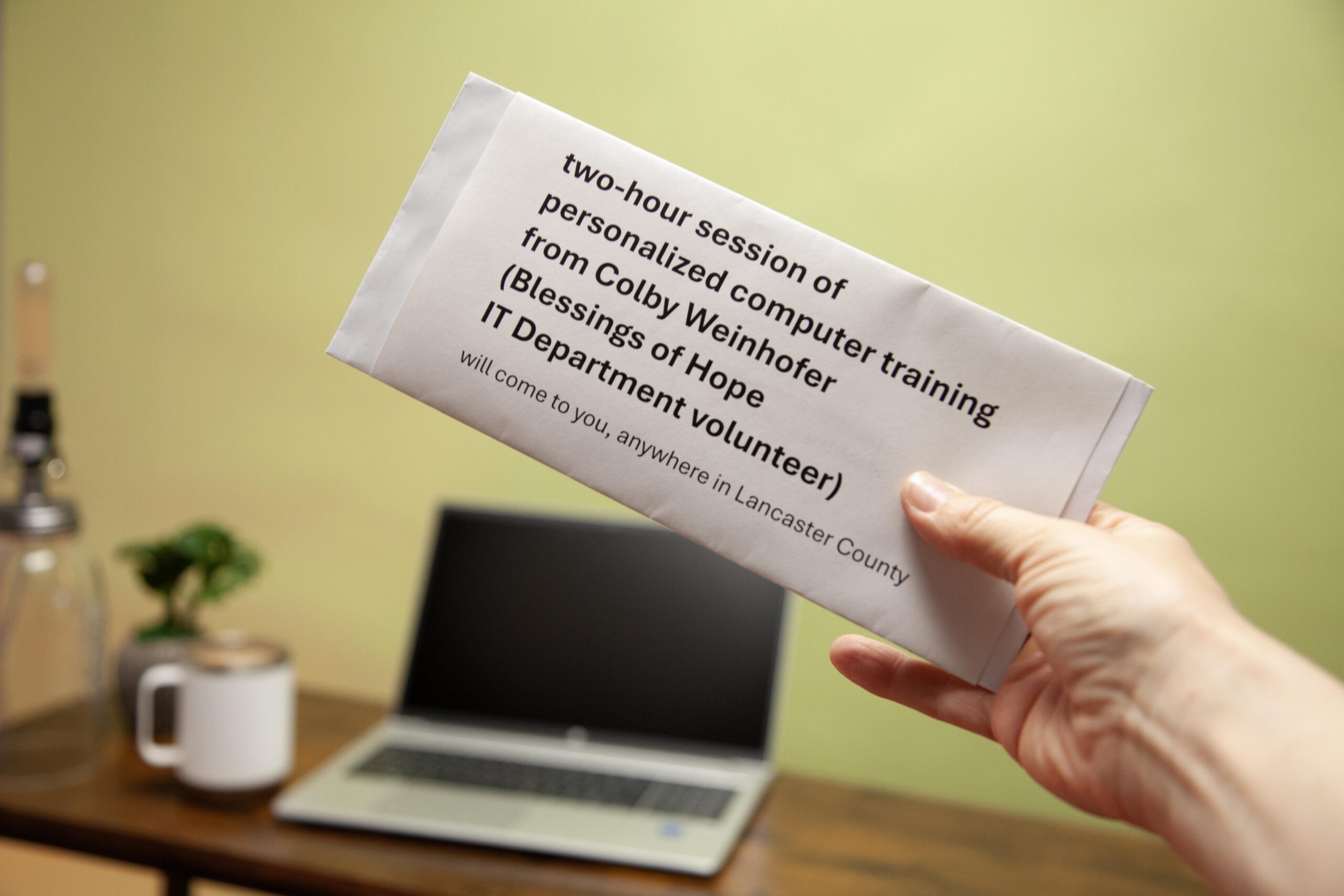 Hand holding an envelope promoting a two-hour personalized computer training session by Colby Weinhofer from Blessings of Hope, available in Lancaster County, featuring a laptop and a coffee cup in the background.