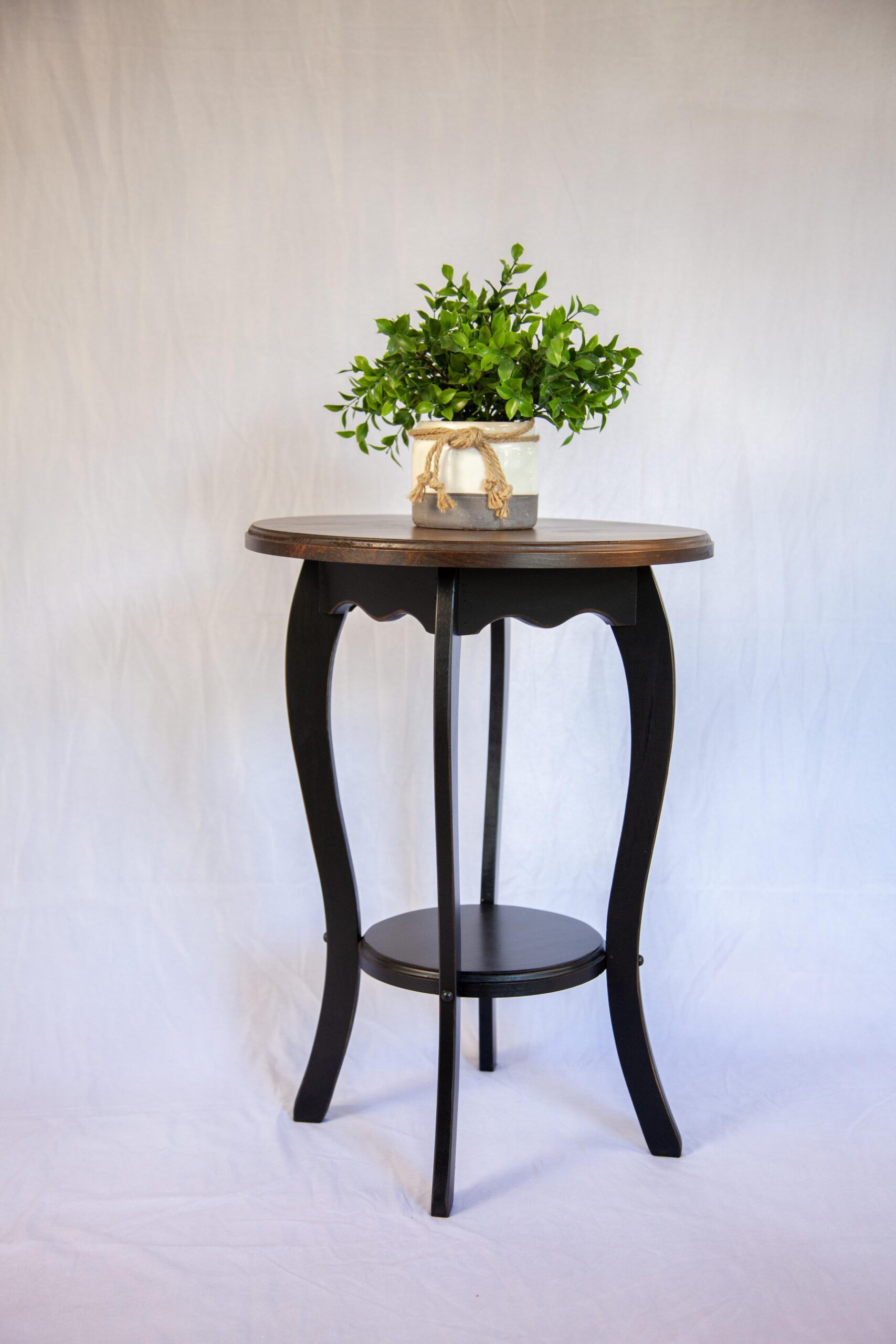 Decorative black side table with a round wood top and curved legs, featuring a potted green plant for a touch of nature against a light background. Ideal for home decor or indoor gardening.