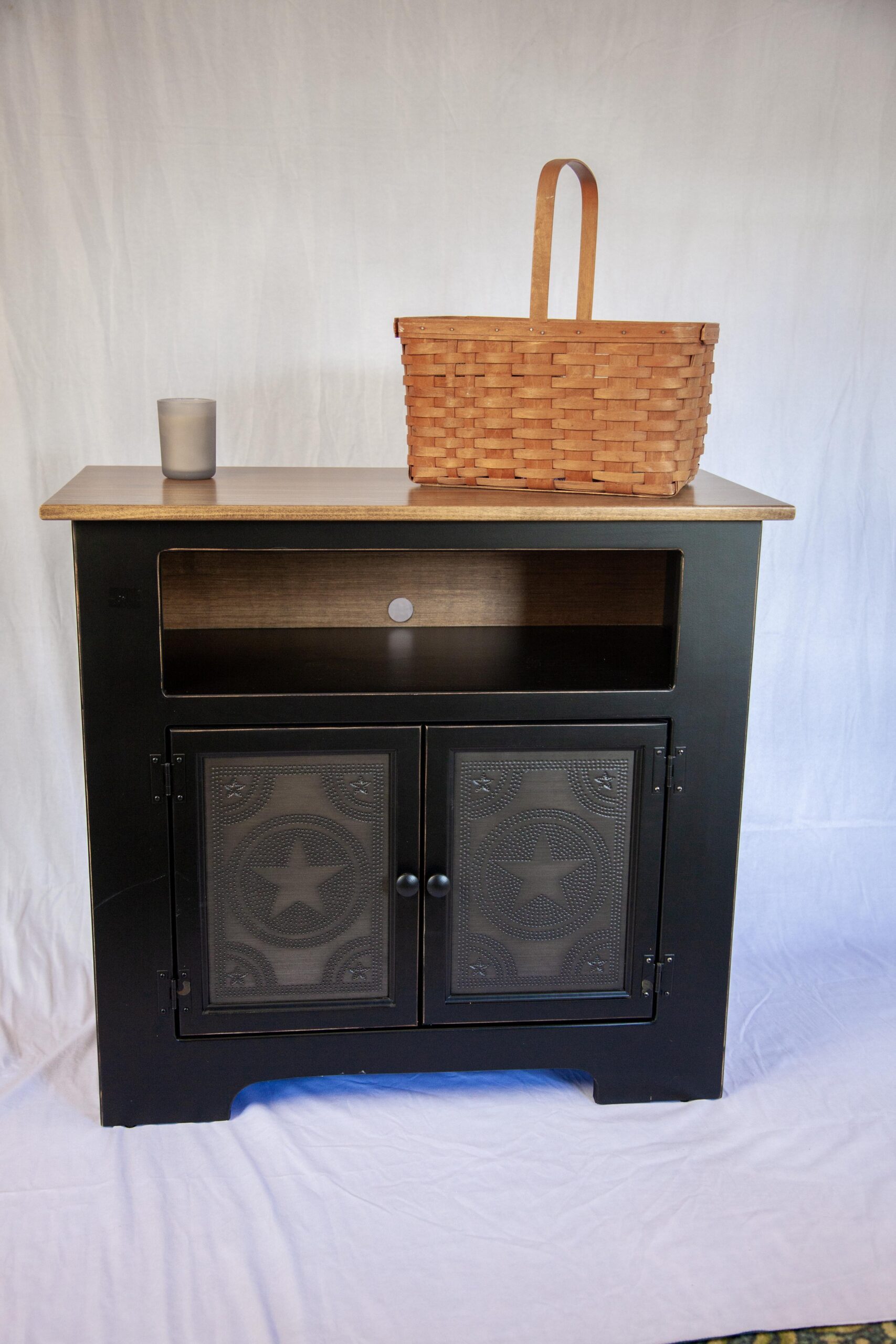 Black cabinet with star-patterned doors, topped with a wicker basket and a glass candle holder against a white backdrop.