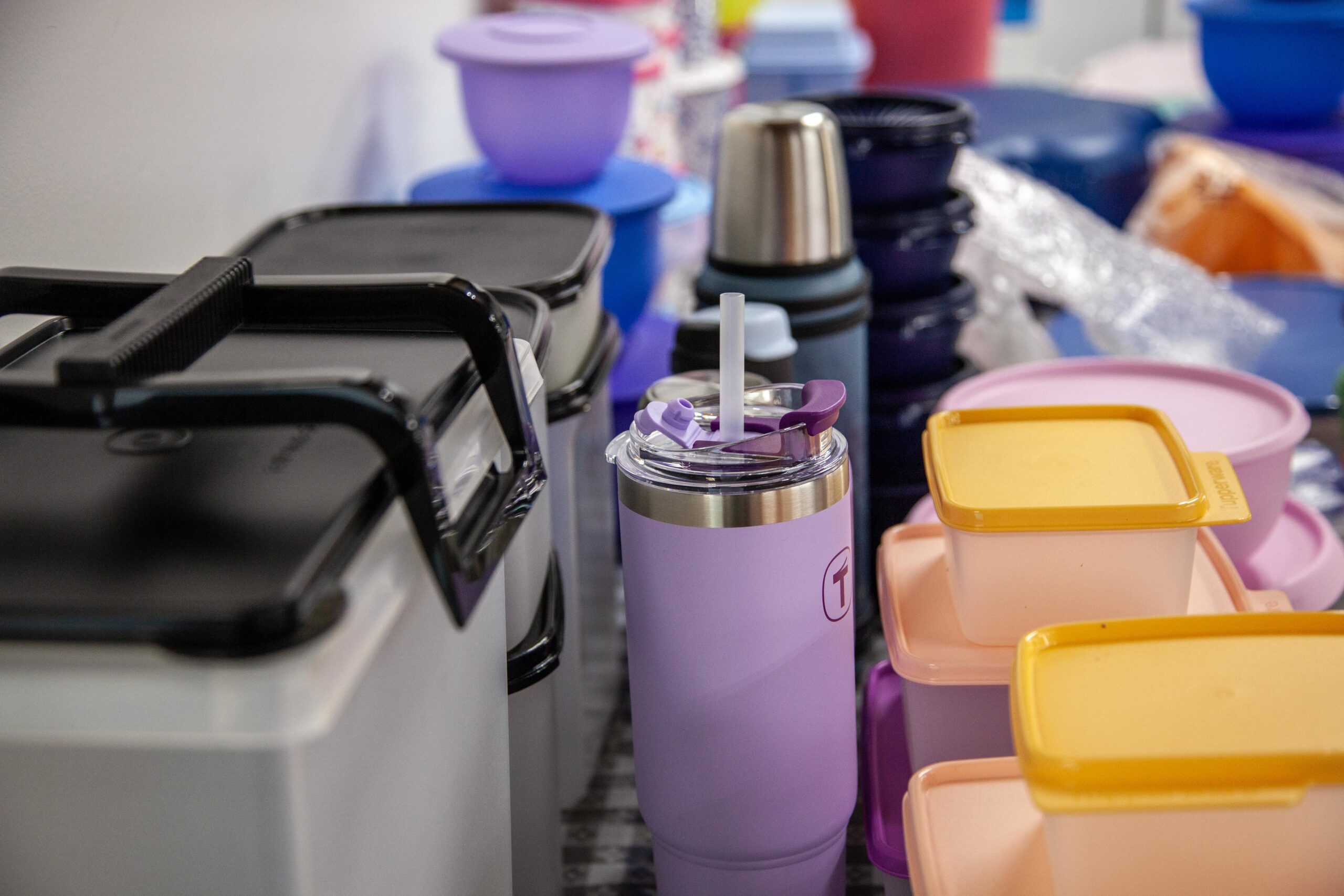 Assorted plastic containers and drinkware displayed on a table, including a purple travel tumbler with a lid and straw, various storage boxes, and colorful bowls, showcasing home organization and kitchen storage solutions.
