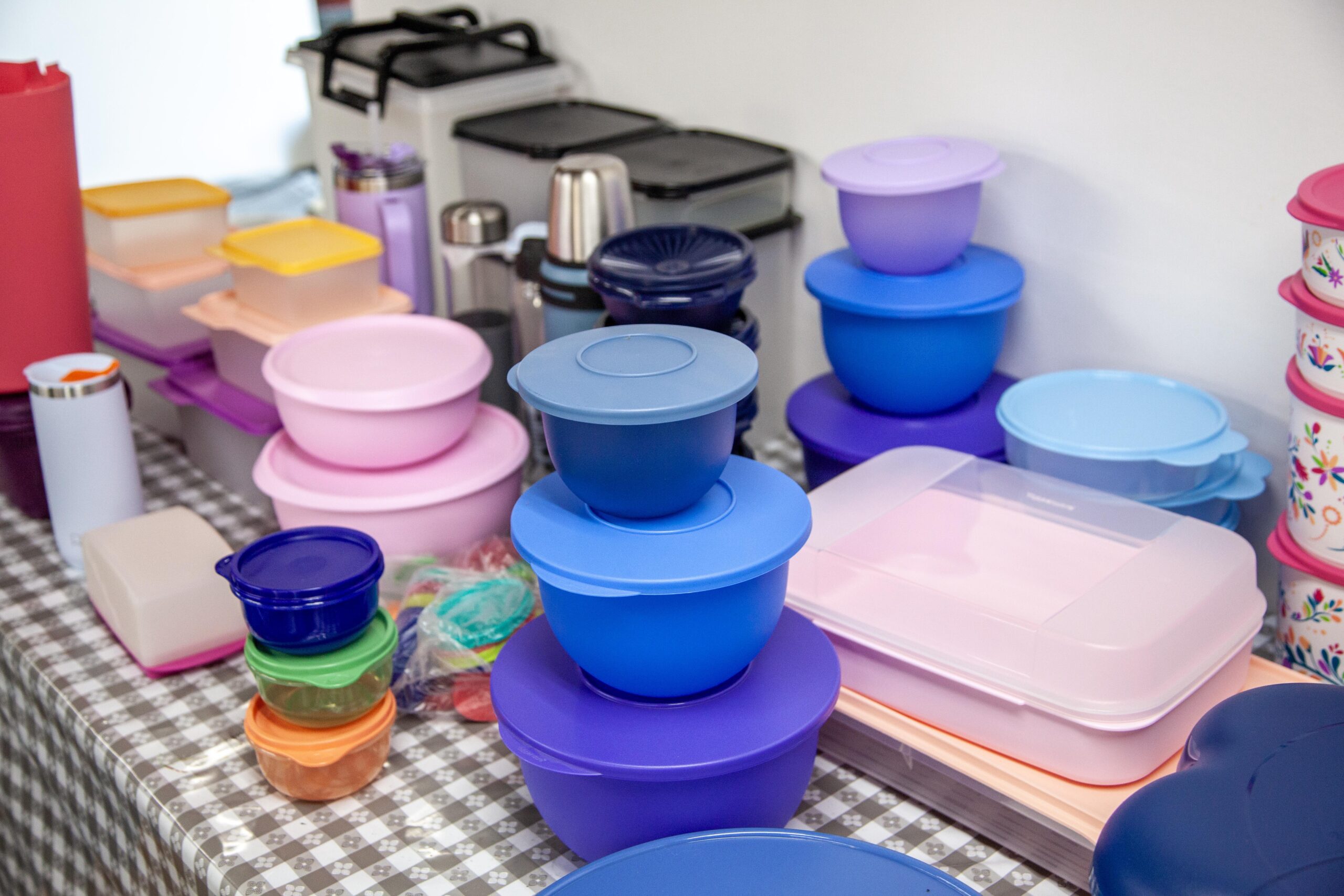 Colorful plastic storage containers and bowls stacked on a table, showcasing various sizes and shapes, with additional kitchen items in the background. Perfect for kitchen organization and food storage solutions.