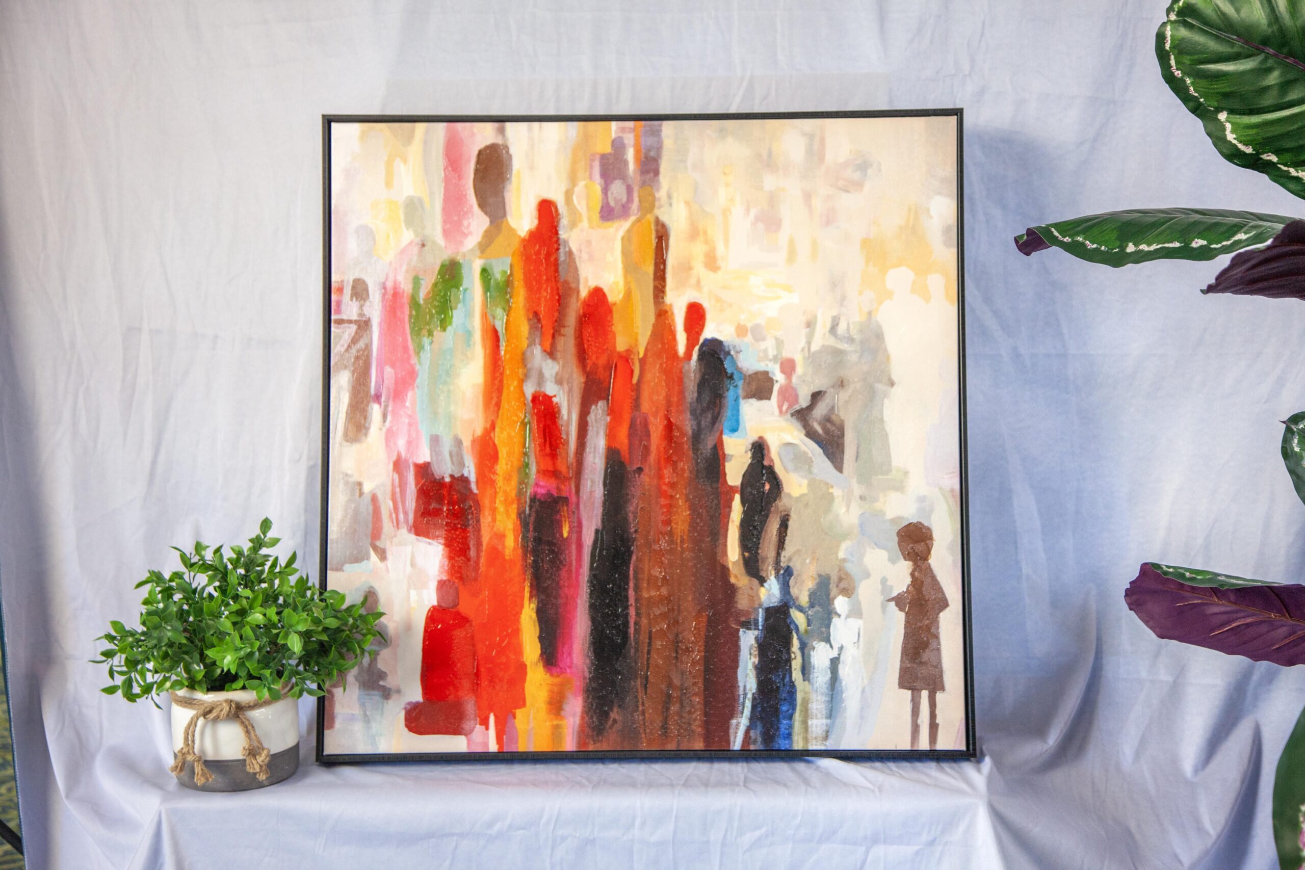 Colorful abstract painting depicting silhouettes of people in vibrant hues, displayed on a white backdrop with a potted plant on the left, creating a modern artistic ambiance.