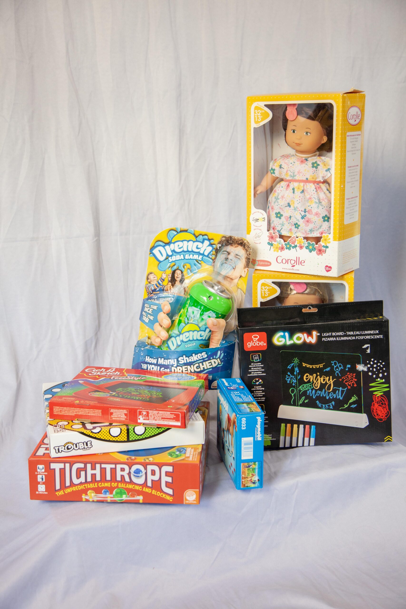 Colorful assortment of children's toys including a Corolle doll, Drench soda game, Glow light board, and various board games like Tightrope and Trouble, arranged on a white backdrop.