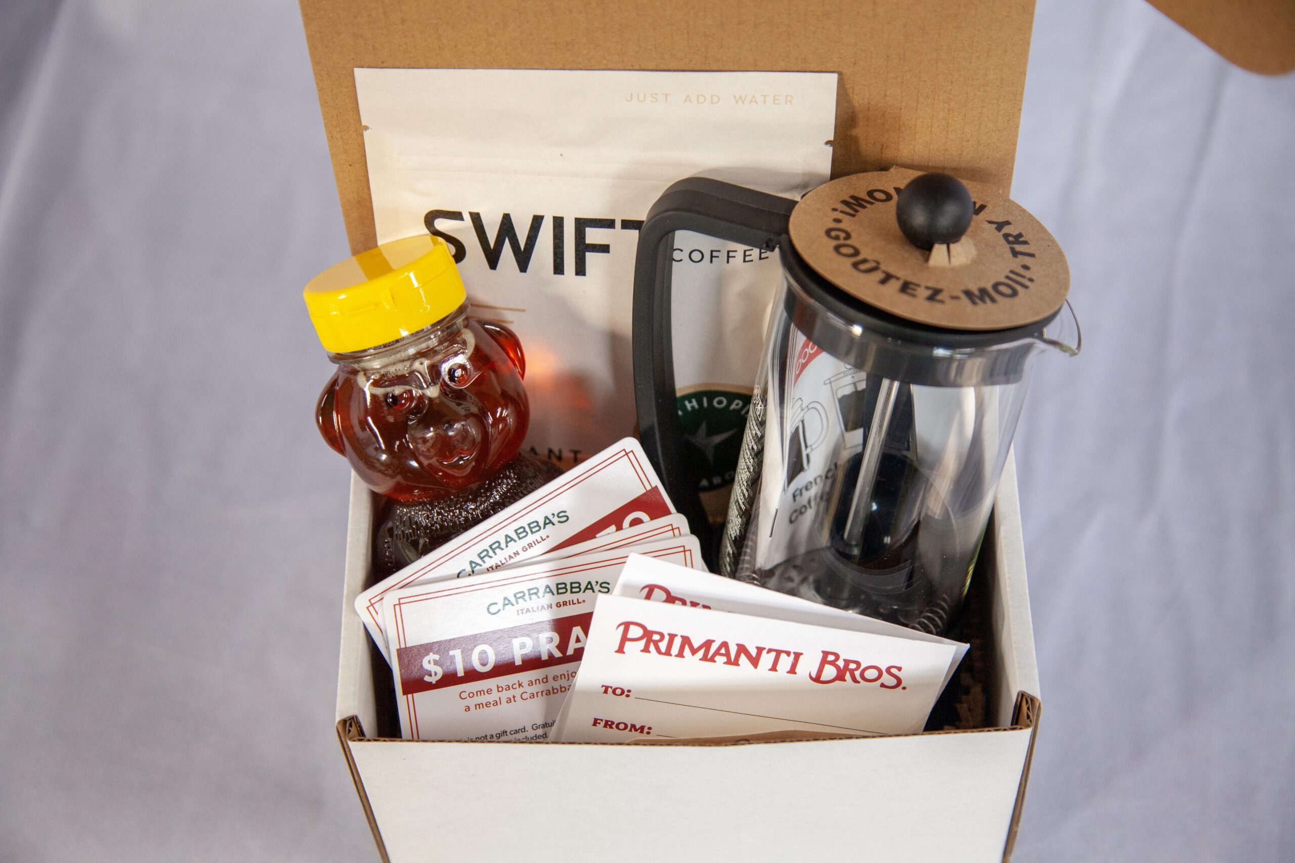Gift box containing coffee essentials, including Swift Coffee, a honey jar, and gift cards from Carrabba's Italian Grill and Primanti Bros, along with a French press for brewing.