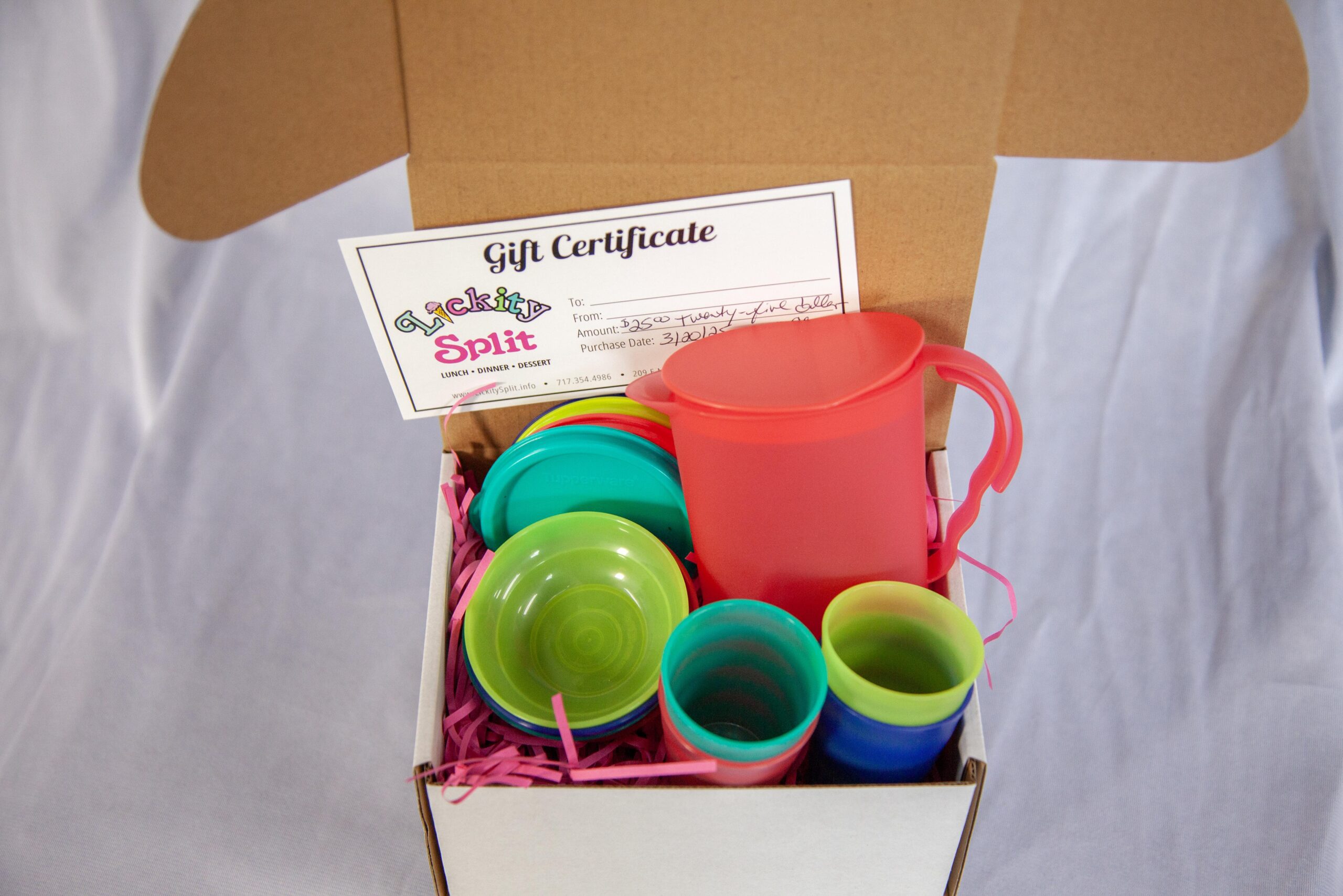 Colorful gift set from Lickity Split featuring a gift certificate, various plastic cups, and a pitcher, arranged in a decorative box filled with shredded paper. Ideal for birthdays or special occasions.