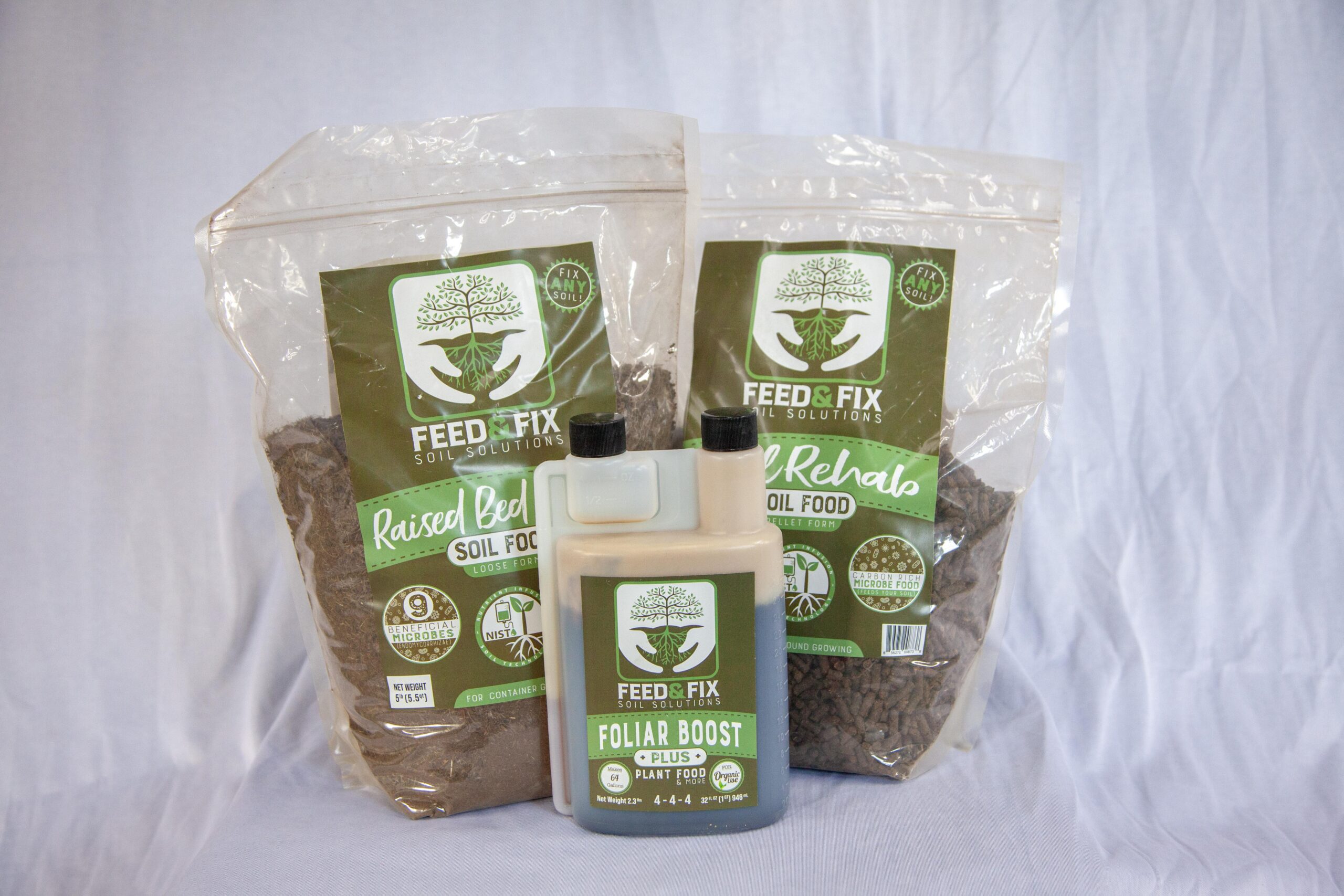 Bags of Feed & Fix soil food products labeled "Raised Bed Soil Food" and "Soil Rehabs" alongside a bottle of "Foliar Boost" plant food, showcasing organic gardening solutions for improving soil health and plant growth.
