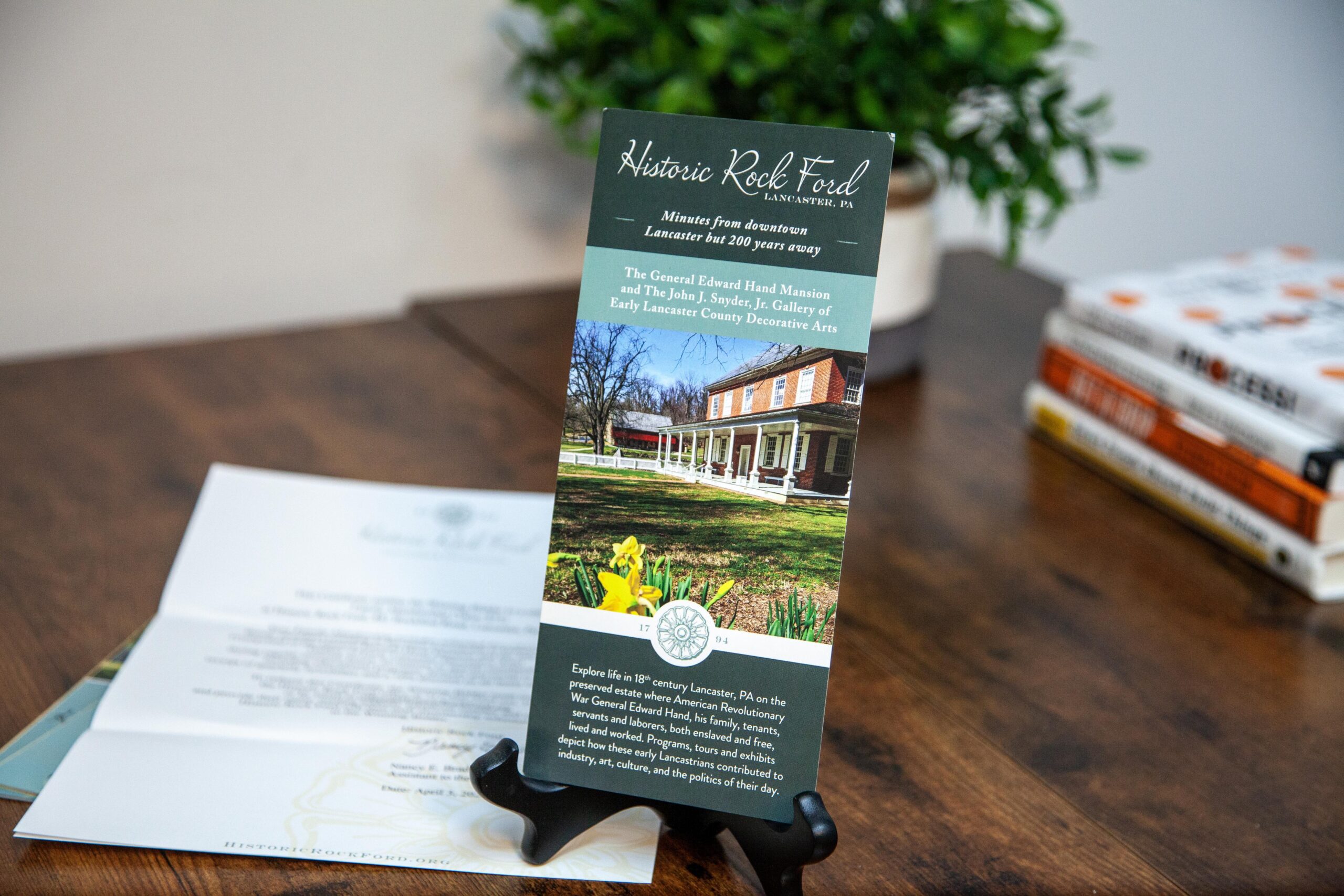 Brochure for Historic Rock Ford in Lancaster, PA, featuring the General Edward Hand Mansion and John J. Snyder, Jr. Gallery. Highlights regional history, events, and exhibits related to 18th-century life in Lancaster County. Displayed on a wooden table with a plant in the background.