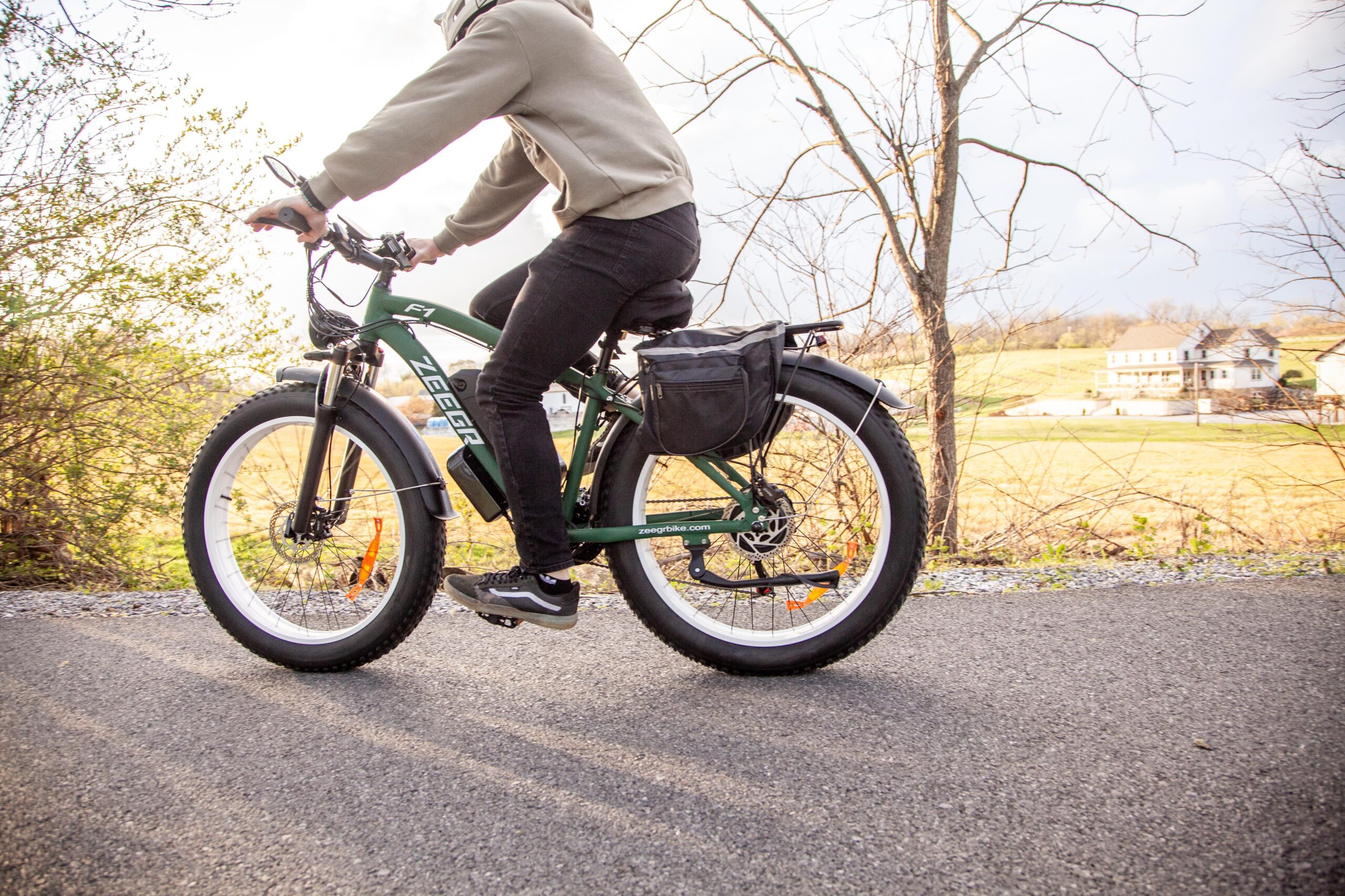 A person riding a green electric bike with wide tires on a paved path, surrounded by bushes and fields, showcasing a scenic rural landscape in the background.