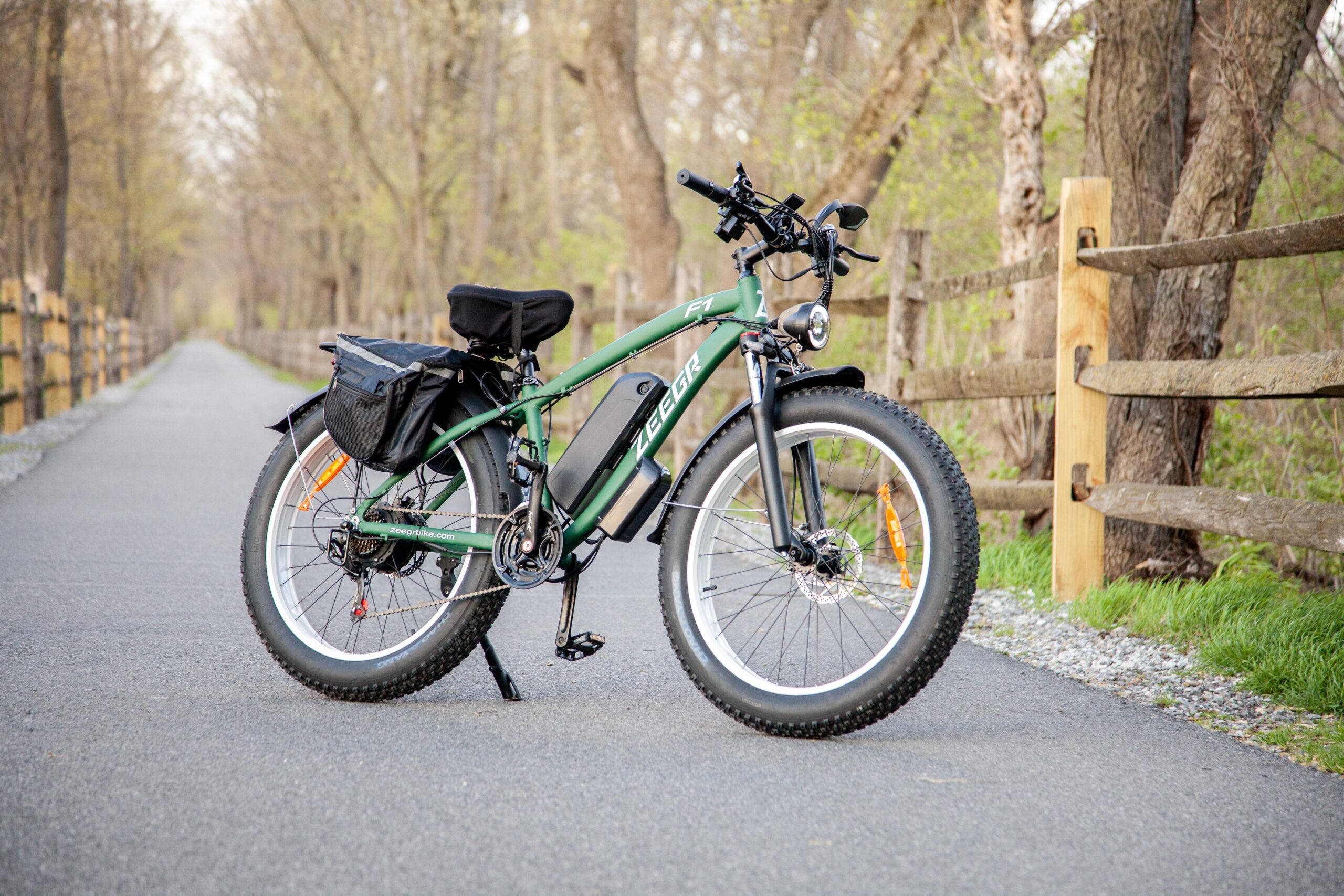 Electric fat bike parked on a scenic trail lined with trees and wooden fencing, showcasing its sleek design and storage bag, perfect for outdoor adventures and commuting.