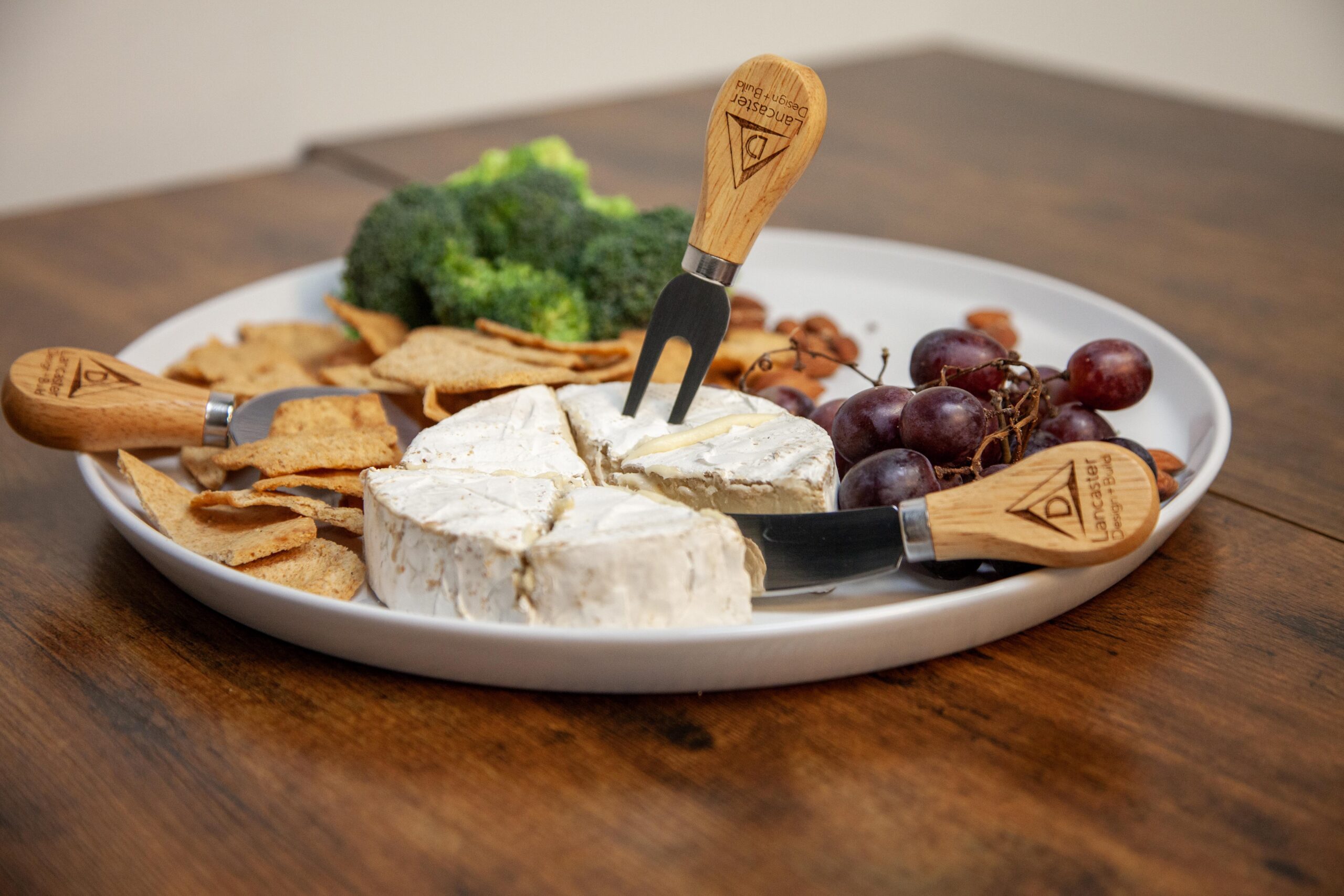 Cheese platter with brie, grapes, broccoli, almonds, and crackers on a white plate with cheese knives.