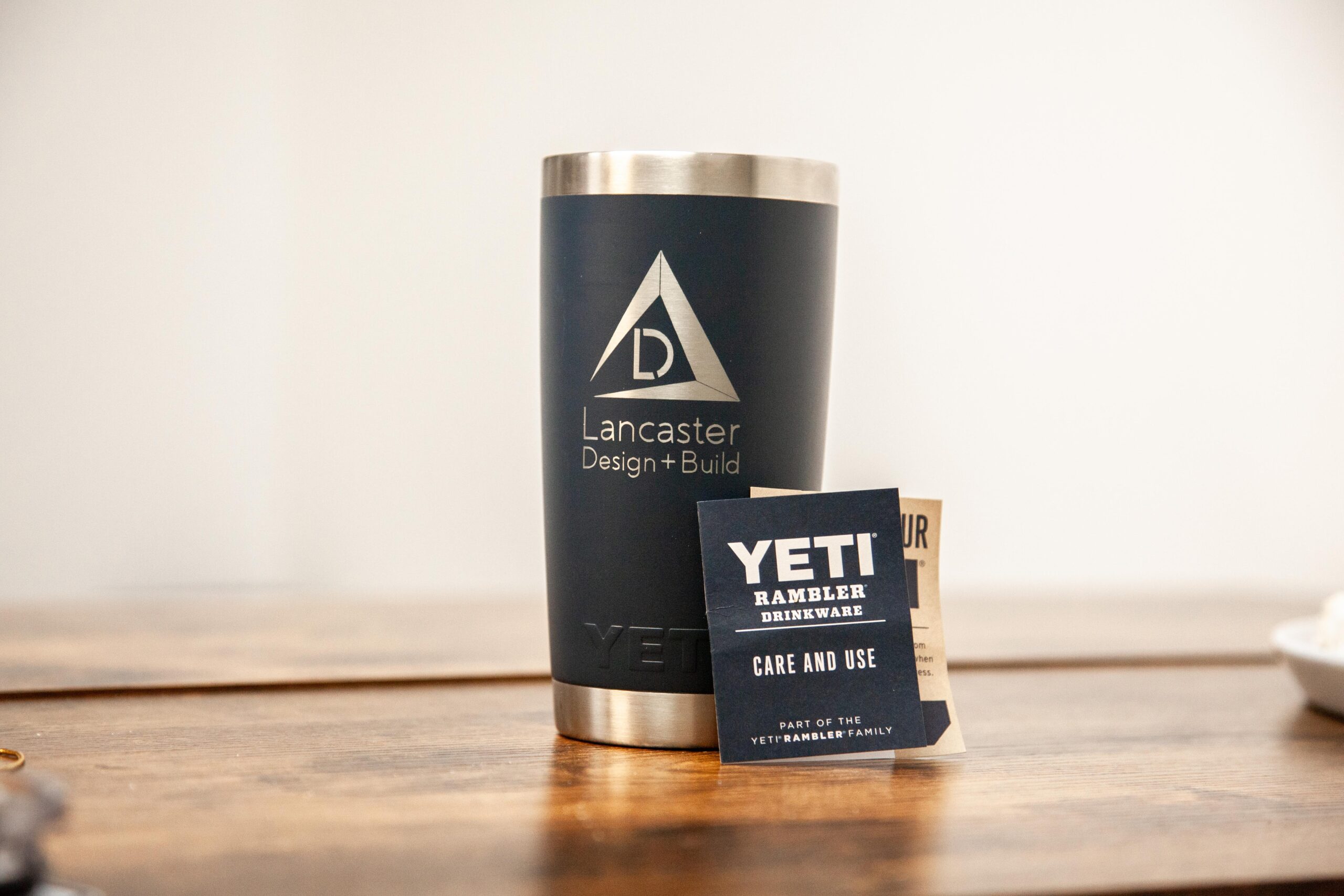 Stainless steel tumbler with Lancaster Design logo and Yeti care instructions card on a wooden surface.