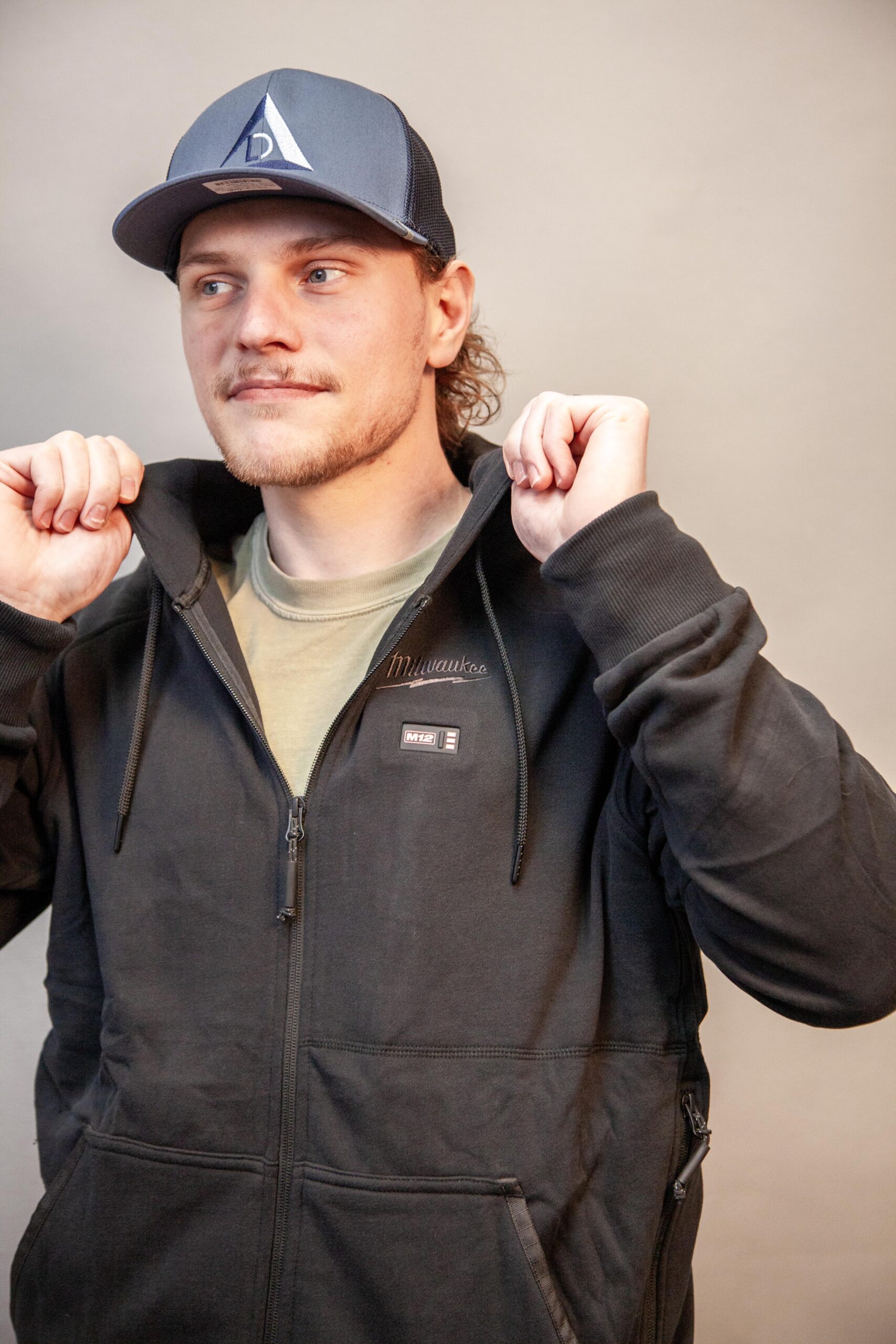 Man wearing a black hoodie and baseball cap, adjusting his collar.