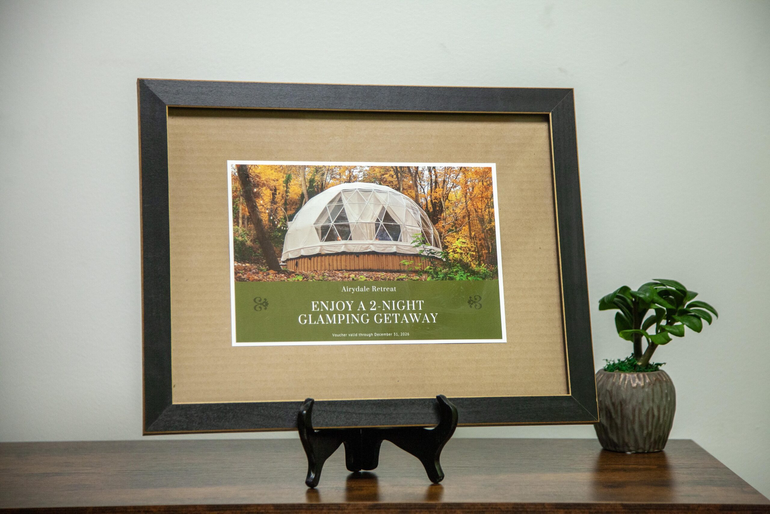 Framed promotional voucher for a two-night glamping getaway at Airydale Retreat, featuring a scenic dome tent surrounded by autumn trees, displayed on a wooden stand beside a small potted plant.