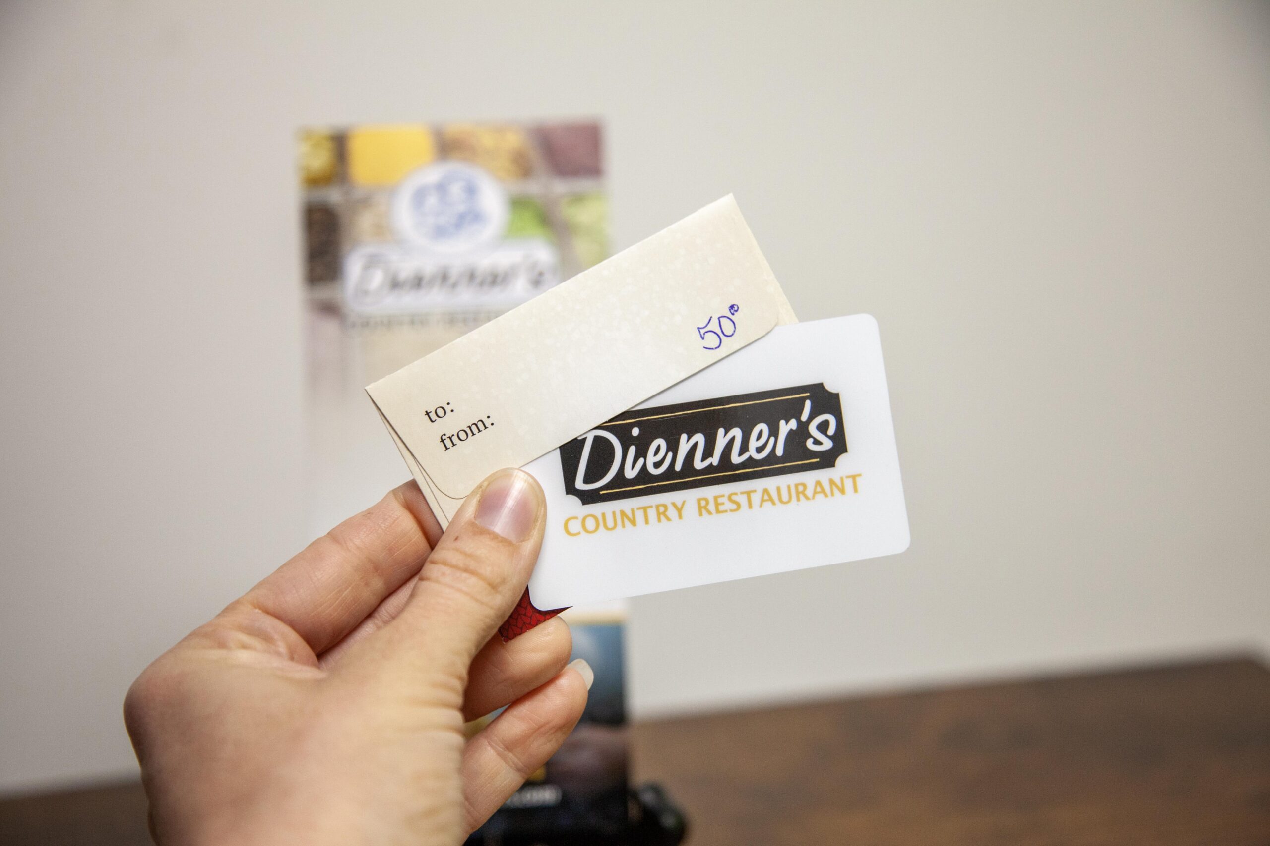 A person holding a Dienner's Country Restaurant gift card and accompanying note card with "to:" and "from:" fields, indicating a thoughtful gift for dining.