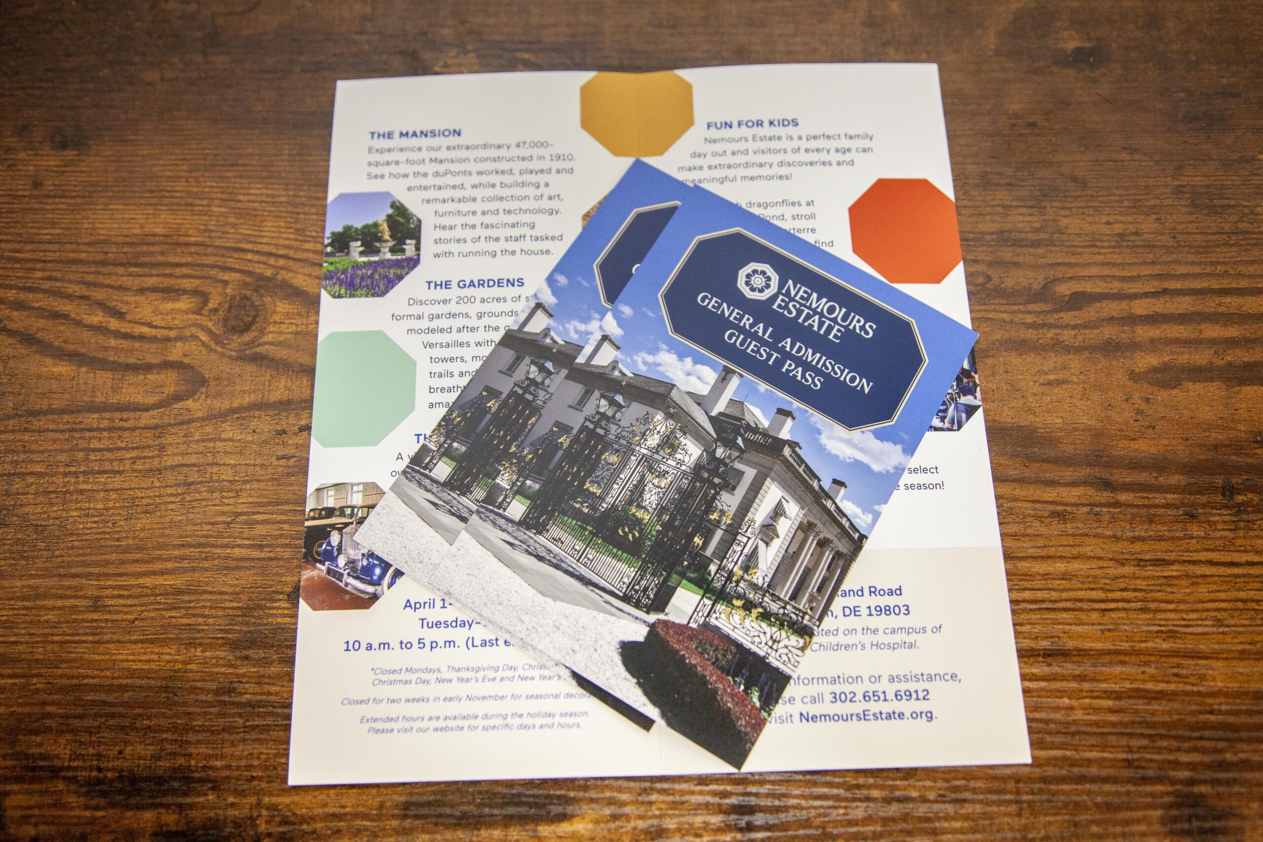 Photo of a guest pass for the Nemours Estate, featuring an image of the estate's entrance, placed over an informational flyer. The flyer includes details about the mansion, gardens, and activities for kids at the estate. The background is a wooden table surface.