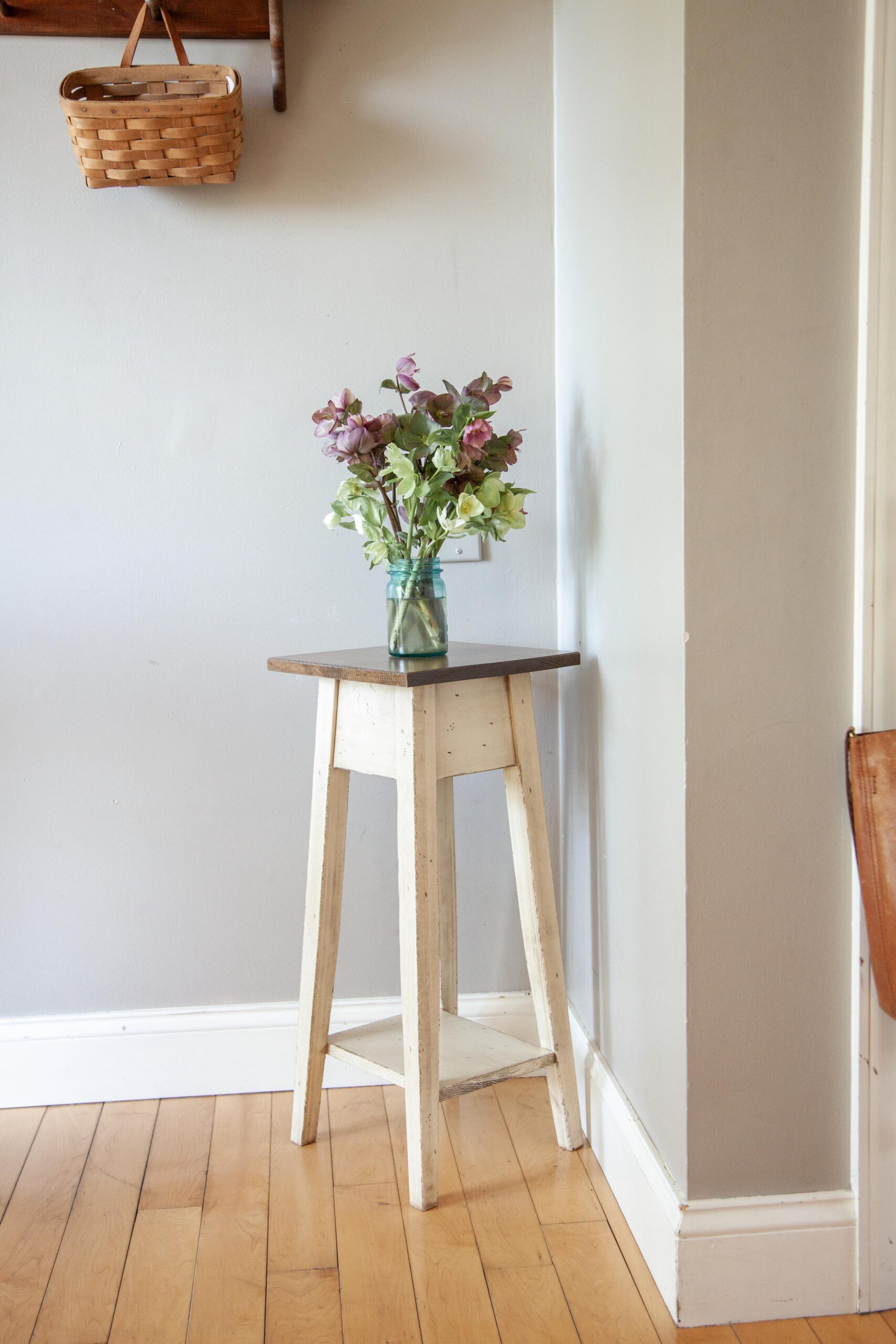 A stylish wooden side table with a dark top, decorated with a vase of fresh flowers, positioned in a cozy corner. A woven basket hangs on the wall in the background against a light gray wall, adding to the inviting ambiance of the room.