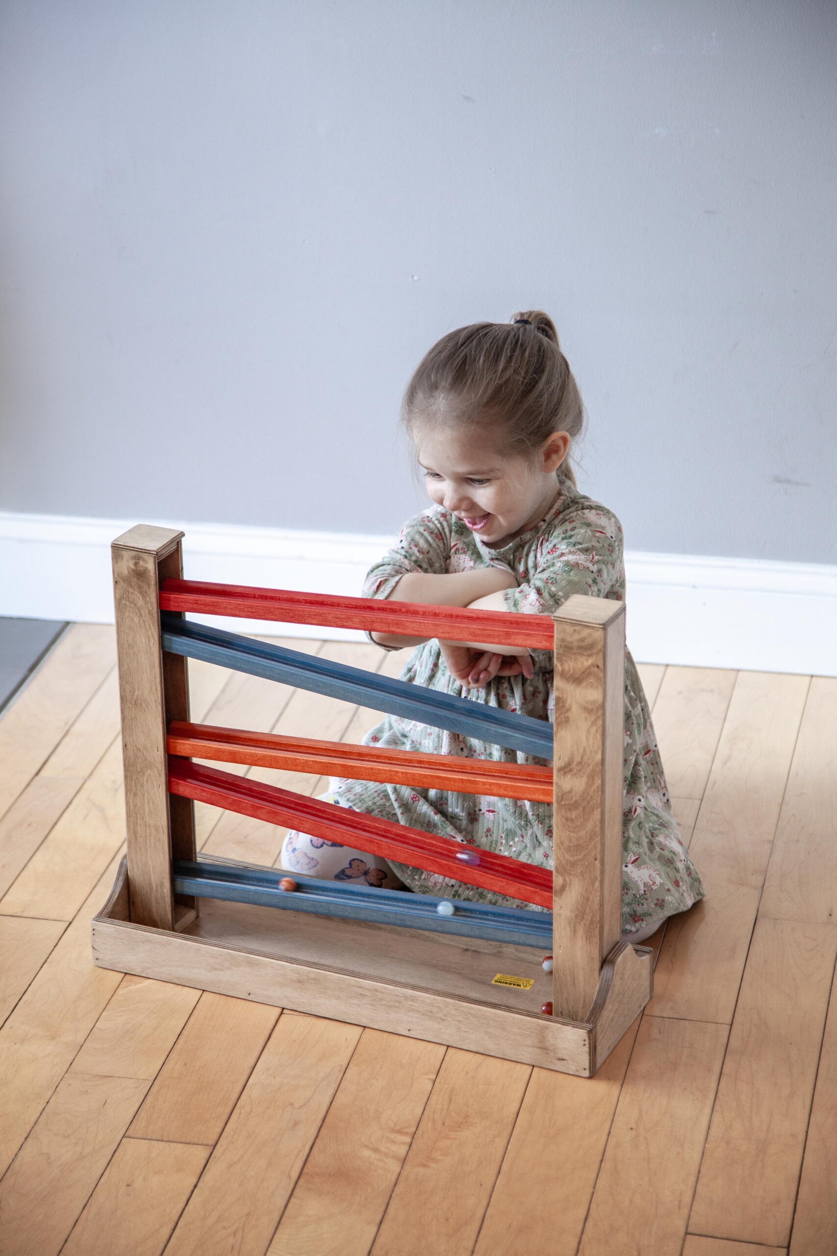 A young girl smiles while playing with a colorful wooden marble run toy made of red, blue, and wooden elements, sitting on a hardwood floor in a bright indoor space.
