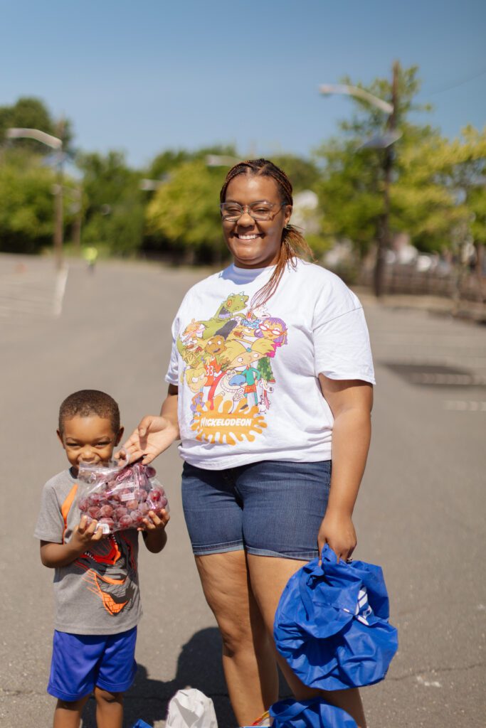 Smiling woman and child holding grapes outdoors on a sunny day, wearing a cartoon t-shirt and shorts.