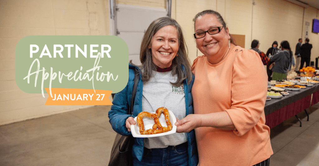 Two women smiling and holding a plate of pretzels at a Partner Appreciation event on January 27, with a buffet table in the background featuring various foods.