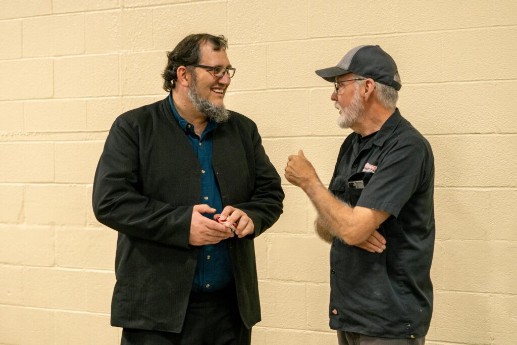 Two men engaged in a friendly conversation, one wearing a black jacket and the other in a dark shirt with a cap, standing against a light-colored wall.