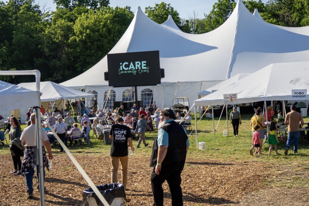 Crowd enjoying the iCARE Community Event under a large tent, with food booths and a big screen displaying event information, surrounded by trees and green grass.