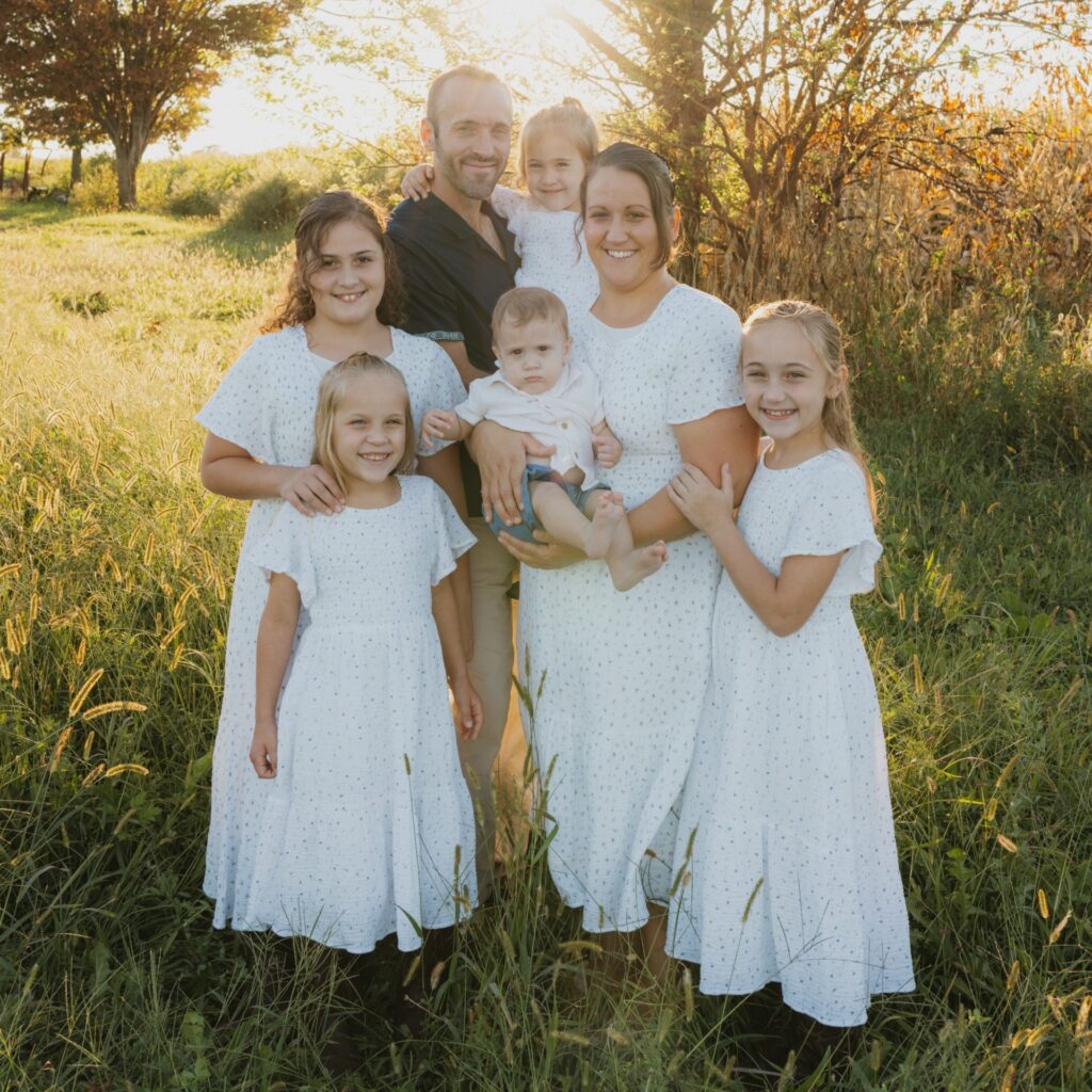 Family portrait in a sunlit field, featuring a man and woman holding a young child, surrounded by five girls in matching white dresses. The scene captures a warm, joyful moment in nature, highlighting family bonds and outdoor beauty.