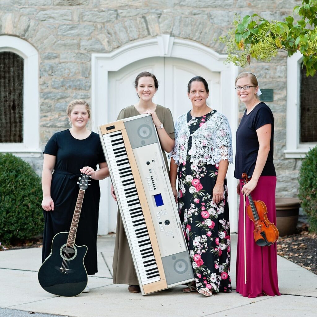 Four women musicians pose outdoors in front of a stone building, showcasing a guitar, keyboard, and violin. They wear elegant dresses and smile, representing a musical group or ensemble ready for a performance.