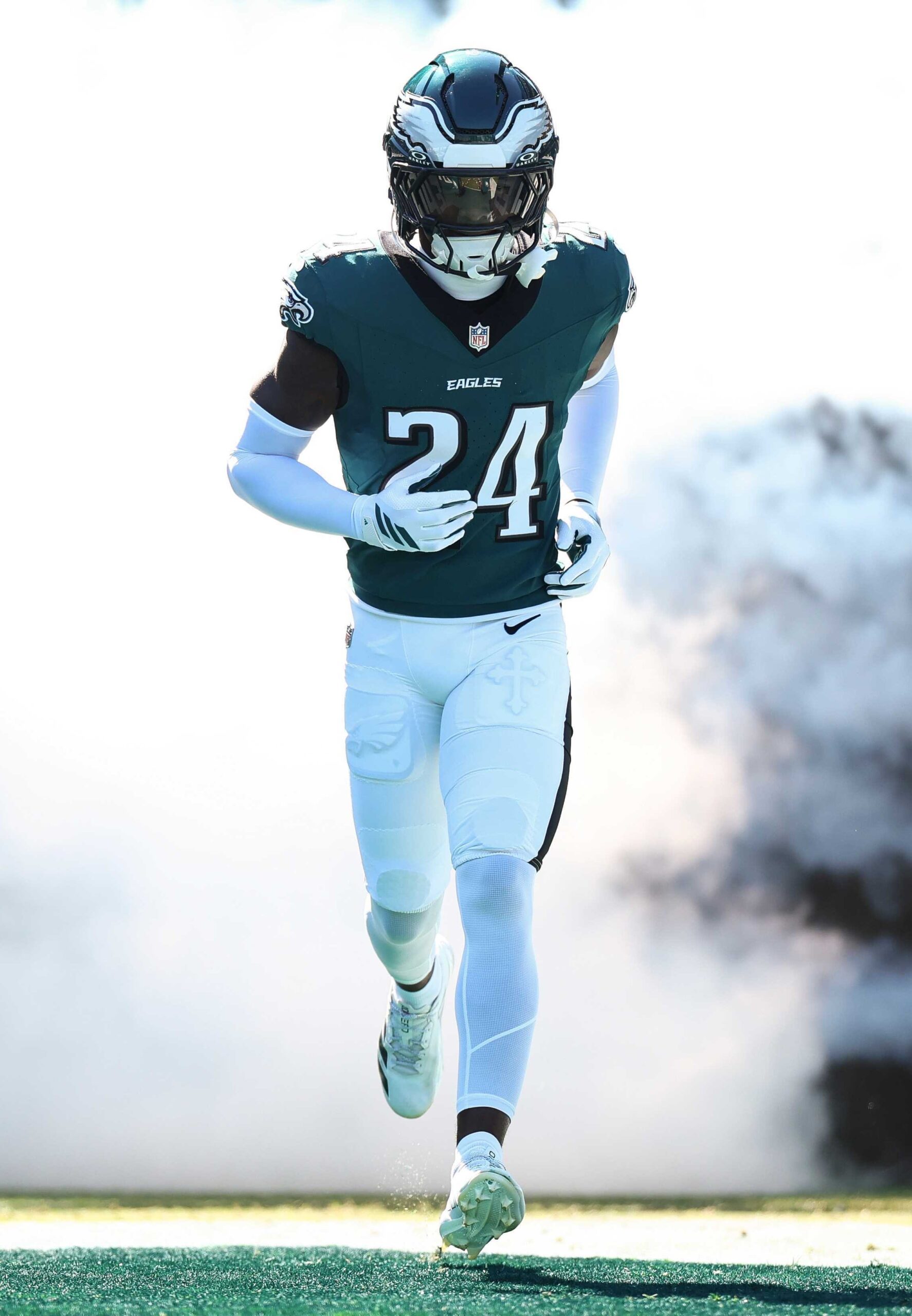 Philadelphia Eagles player in a teal and white uniform, wearing number 24, running onto the field through a cloud of smoke during a game. The player is equipped with a helmet and gloves, showcasing the team's branding.