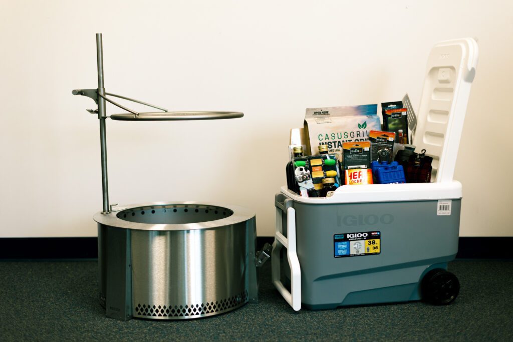 Stainless steel fire pit with a cooking grate next to an Igloo cooler filled with grilling supplies and outdoor gear, set against a plain background.